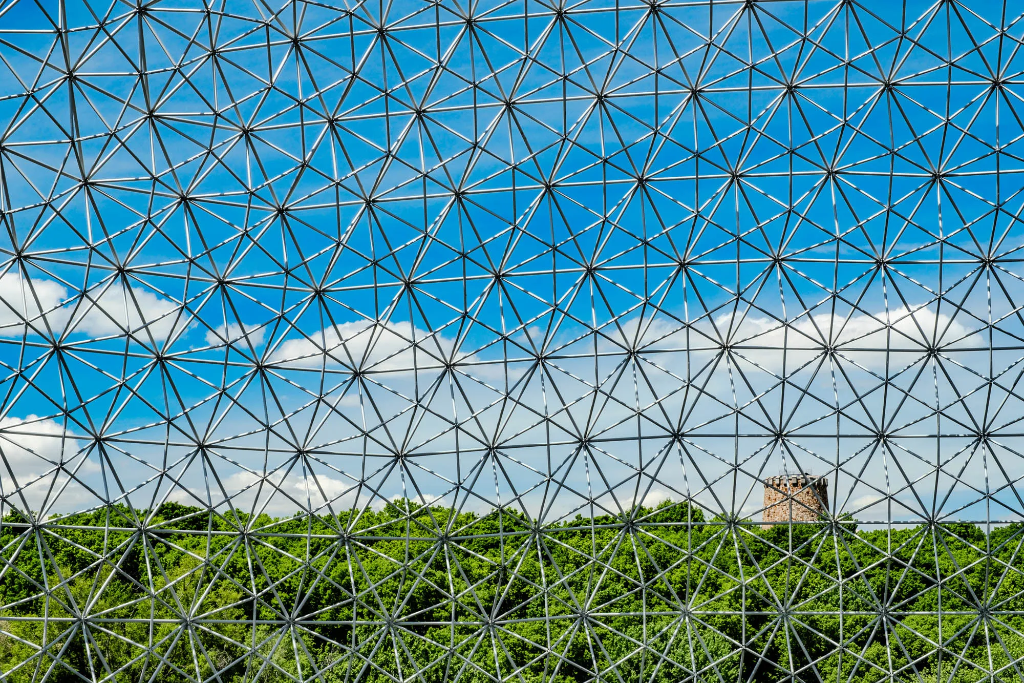 The image shows a close-up view of a geodesic dome. The dome is made of a series of interconnected struts that create a repeating pattern of triangles. The struts are a light gray color, and they are set against a background of a bright blue sky with white clouds.  Through the dome, you can see a forest of green trees below, with a tall stone tower peeking through the trees in the center. 
