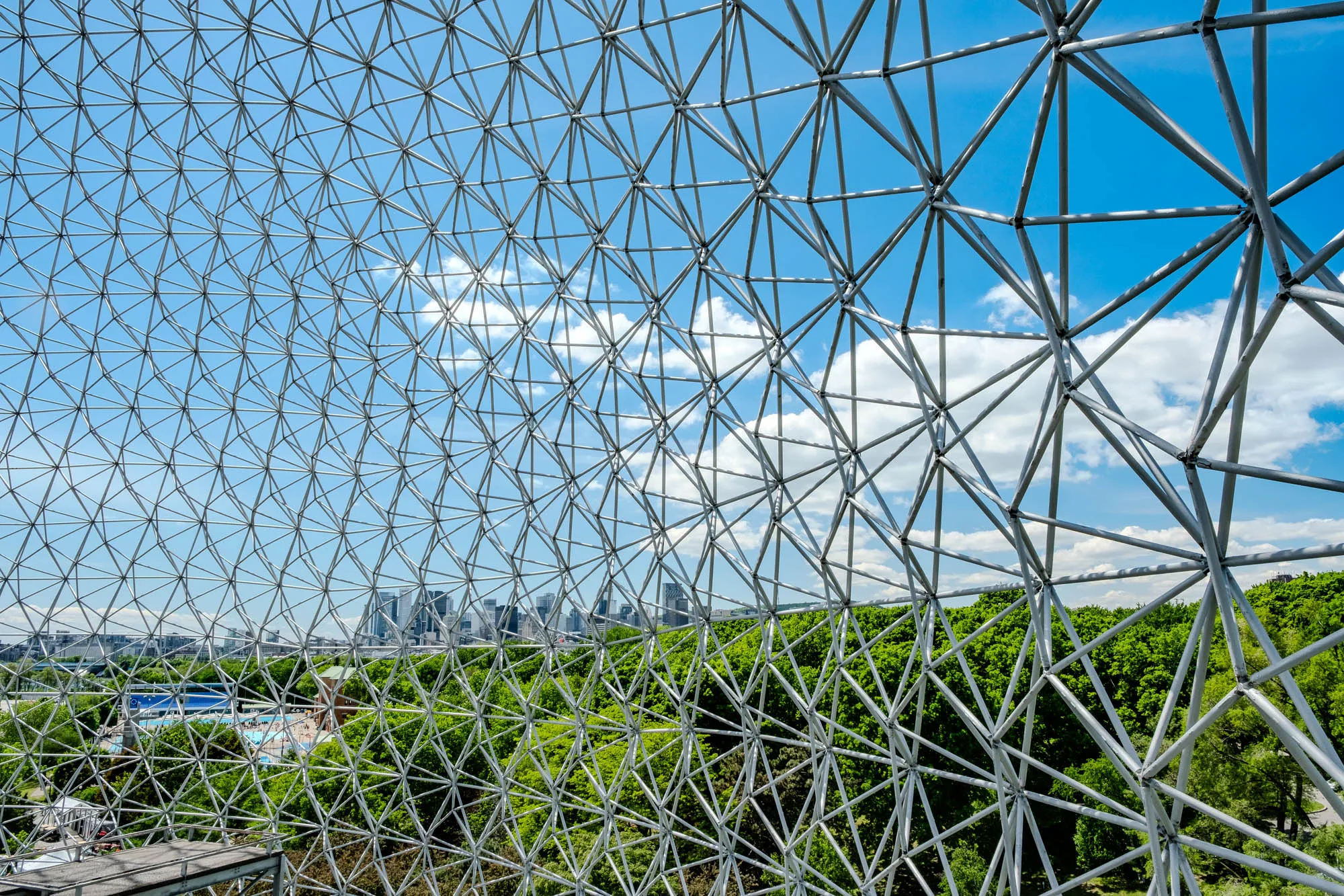 The image shows a close-up of a geodesic dome made of metal bars arranged in a complex network of triangles. The dome is transparent, allowing a view of the sky and the surrounding landscape. The sky is a bright blue with white clouds, and the ground is covered in lush green trees.  In the distance, through the metal framework, a city skyline can be seen, with tall buildings that look like thin rectangles against the blue sky. There is also a hint of a body of water in the foreground, possibly a pond or a pool.  The overall effect is one of a light and airy space, with a sense of openness and perspective.