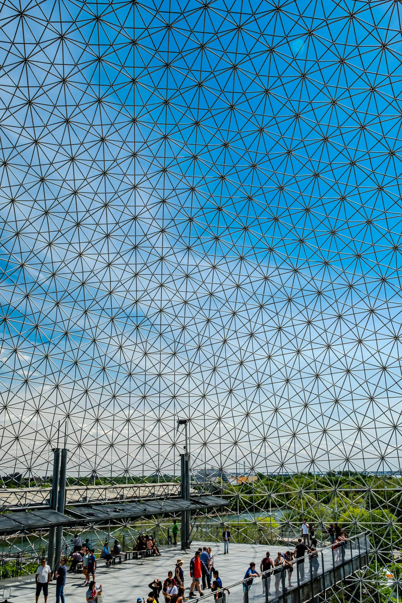 The image shows a view through a geodesic dome. The dome is made of metal struts that form a geometric pattern. The view through the dome shows a lush green forest with a stone tower in the distance. The sky is a bright blue with a few white clouds. The scene is bright and sunny.