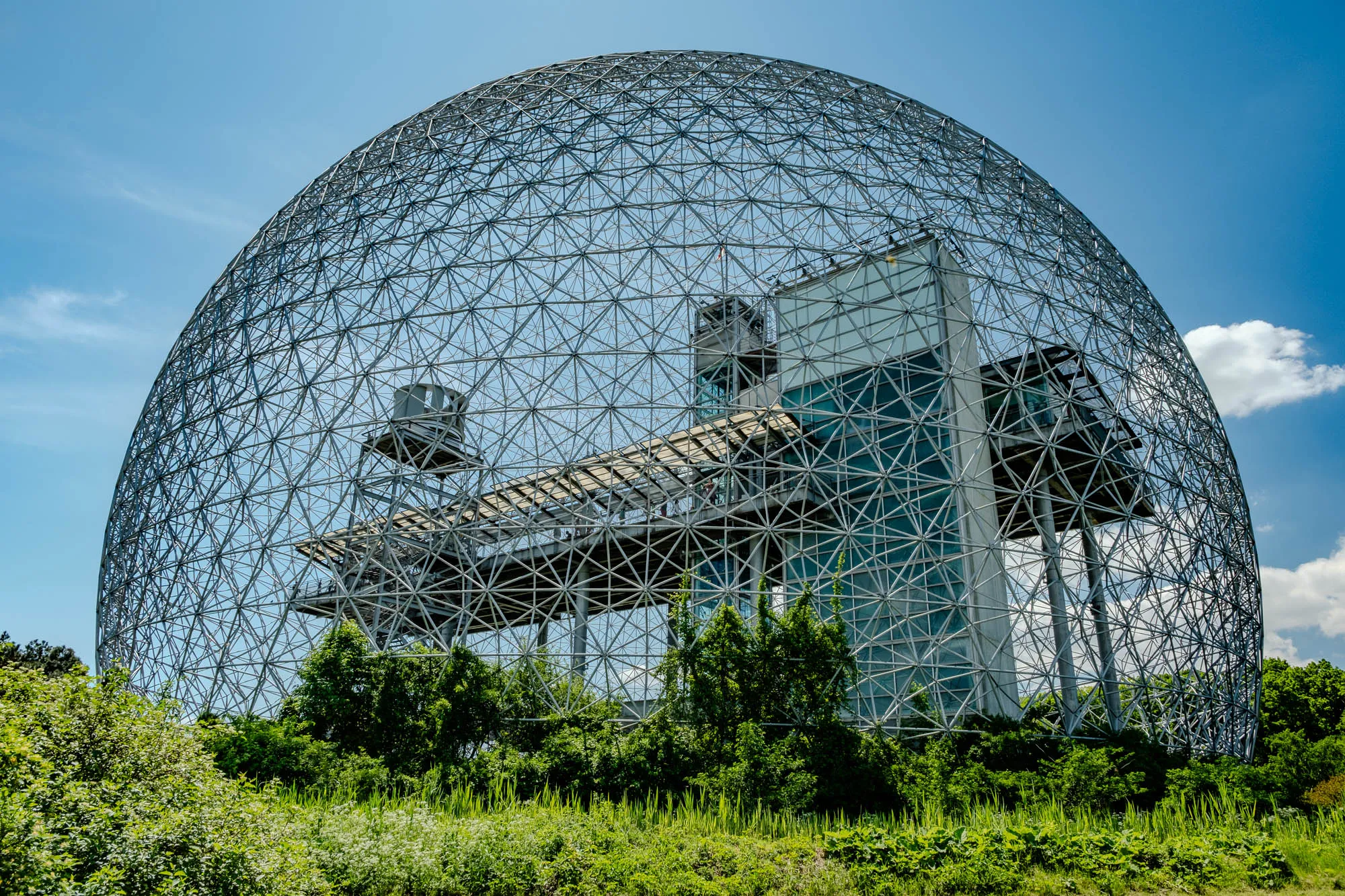 The image shows a large, metal geodesic dome structure. The dome is made up of a grid of interconnected beams, creating a network of triangles that cover the entire surface. The dome is partially obscured by a modern building with glass windows. The building is partially visible in the lower right corner. The dome is surrounded by trees and foliage. The sky is clear with a few clouds in the background.