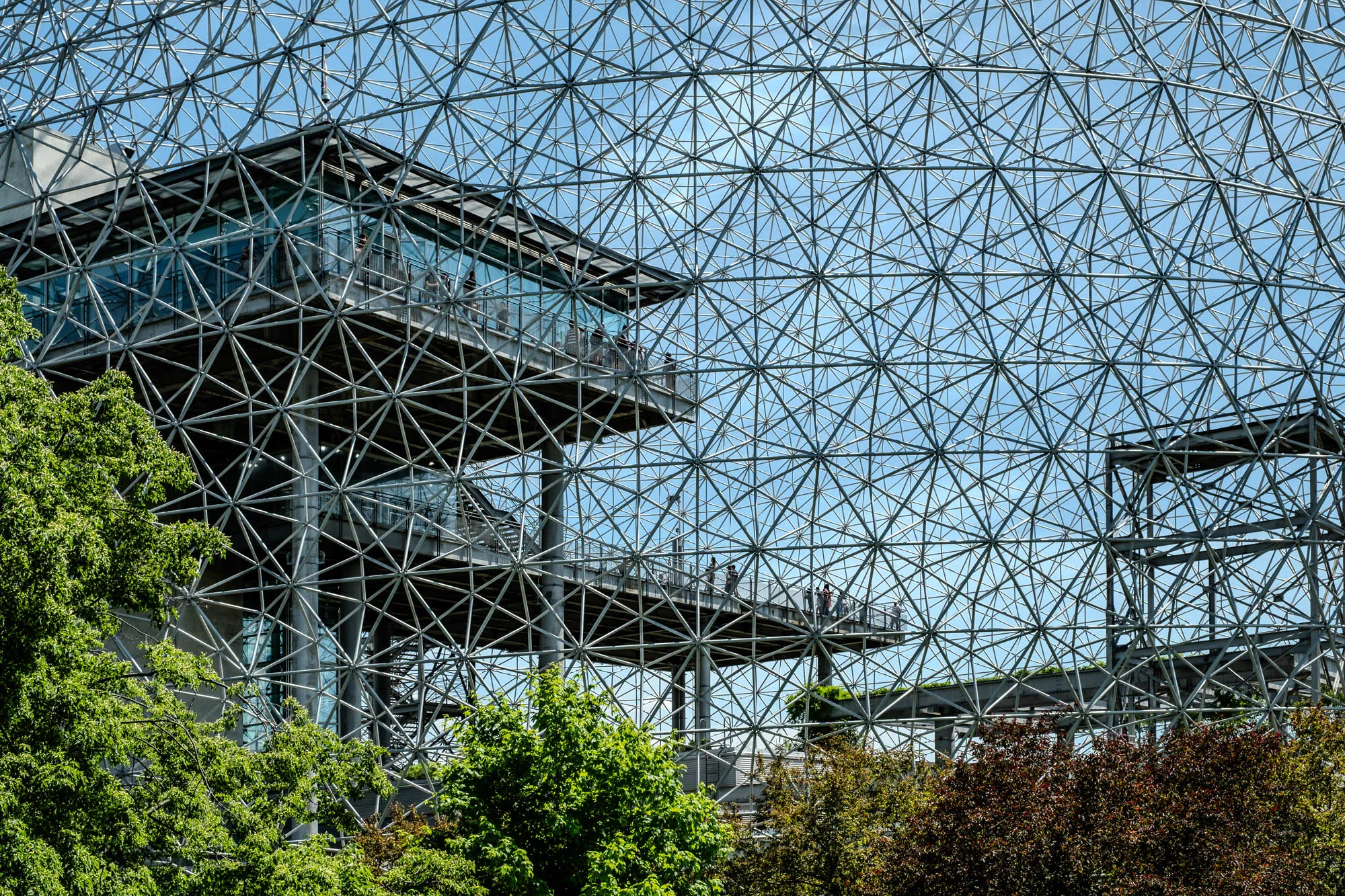 A low-angle view of a large, metal geodesic dome with a platform and railings near the top. The dome is made up of a lattice of steel beams, and the sky can be seen through the open spaces. There are a few people standing on the platform, and a few trees can be seen at the bottom of the image. The image appears to have been taken in the middle of a sunny day with a bright blue sky.