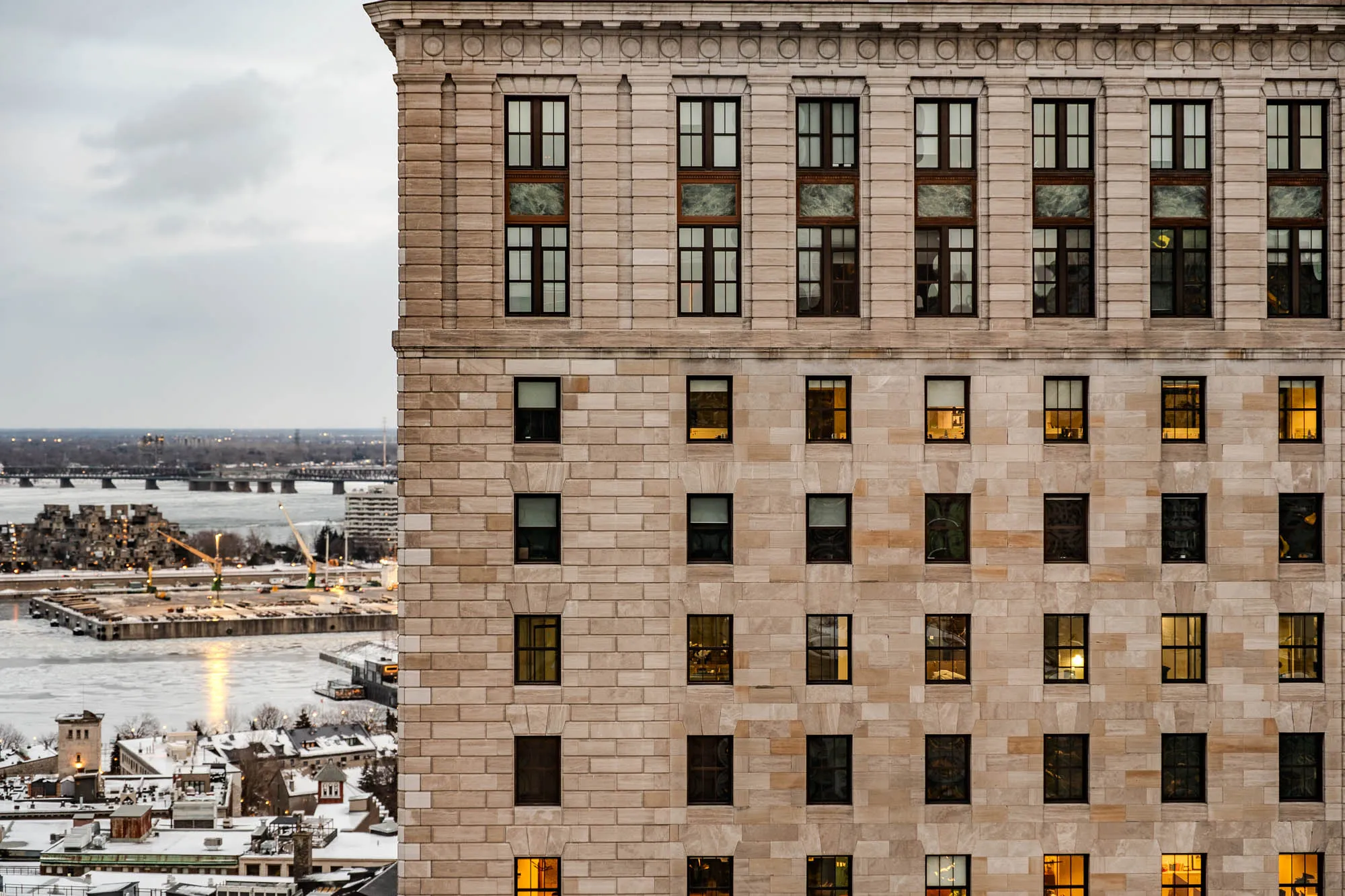 A grand, light-colored stone building dominates the right two-thirds of the frame, its ornate facade featuring rows of rectangular windows. Many windows are dark, reflecting the overcast sky, while others emanate a warm, golden light from within. To the left, a vast, partially frozen river stretches under a muted sky, reflecting bright light patches on its icy surface. Beyond the river, a distinctive cluster of cubist-like residential buildings and distant bridges punctuate the horizon of a snow-covered urban landscape, establishing a cold, early morning scene.