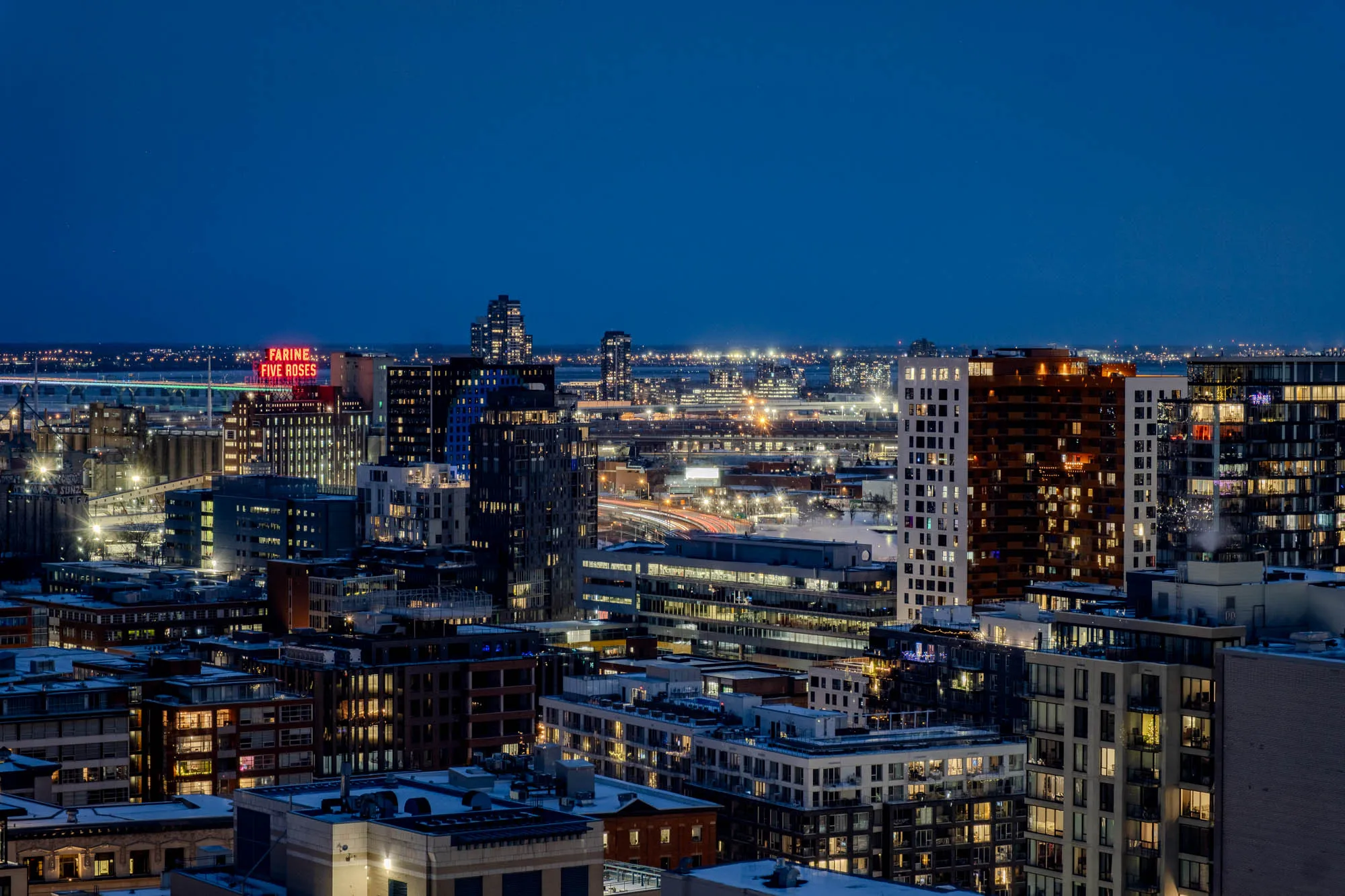 A dense twilight cityscape of Montreal spreads beneath a deep blue sky. In the mid-ground, the iconic red neon sign spelling "FARINE FIVE ROSES" illuminates the industrial buildings near a wide body of water. Streaks of red and white light from vehicles form luminous trails across elevated roadways, a result of the photograph's long exposure. Hundreds of windows in diverse buildings, from older industrial structures to modern high-rises, glow with warm interior light, contrasting with the cool evening tones.