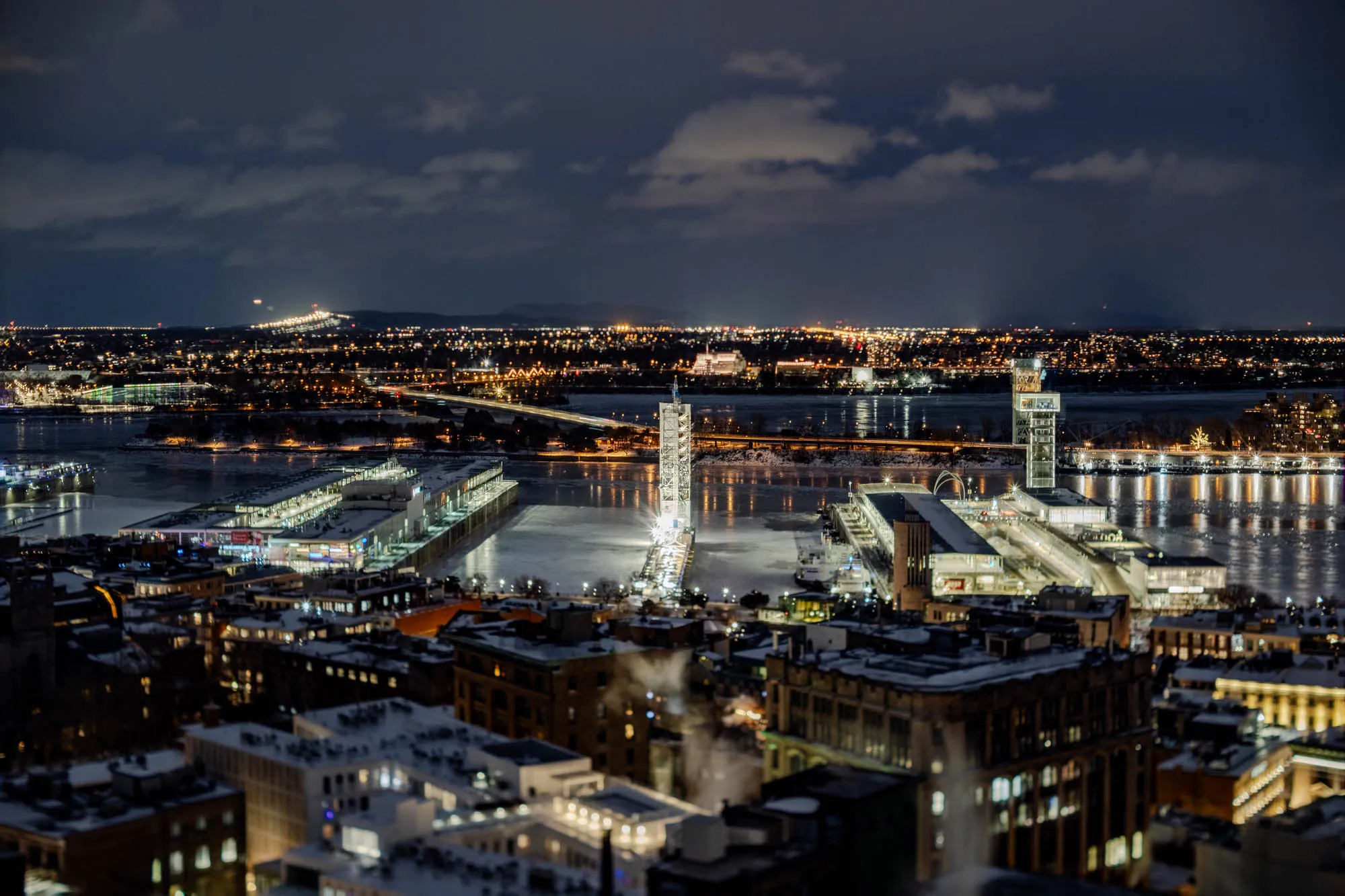 An aerial nighttime vista reveals the Old Port of Montreal, with the partially frozen Saint Lawrence River stretching across the middle ground under a dark, cloudy sky. Two tall, illuminated white structures, including a prominent clock tower and a modern observation tower, punctuate the waterfront, flanked by brightly lit buildings and docks. Long exposure photography renders vehicle headlights and taillights as glowing streaks across distant bridges, creating ethereal lines over the urban landscape. Snow-dusted rooftops in the foreground add texture, while the distant city lights on the horizon glow softly against rolling hills.