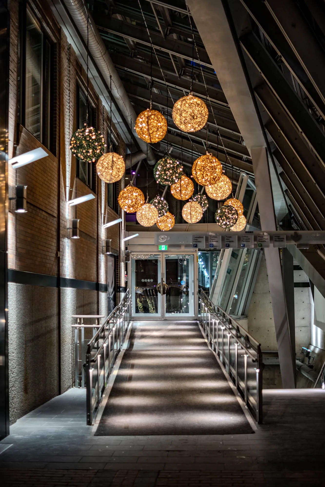 An illuminated architectural walkway ascends towards a glass double door, lined by stainless steel handrails and bathed in the soft glow of numerous spherical pendant lights. A cluster of woven spherical lights, primarily warm amber and some darker green, hangs from the high ceiling, casting dappled patterns onto the dark ramp below. A textured brick wall with horizontal light fixtures defines the left side, contrasting with the angular concrete beams and glass panels forming the right wall and ceiling structure. The wide aperture creates a delicate softness around the ambient details, emphasizing the inviting, festive path ahead.