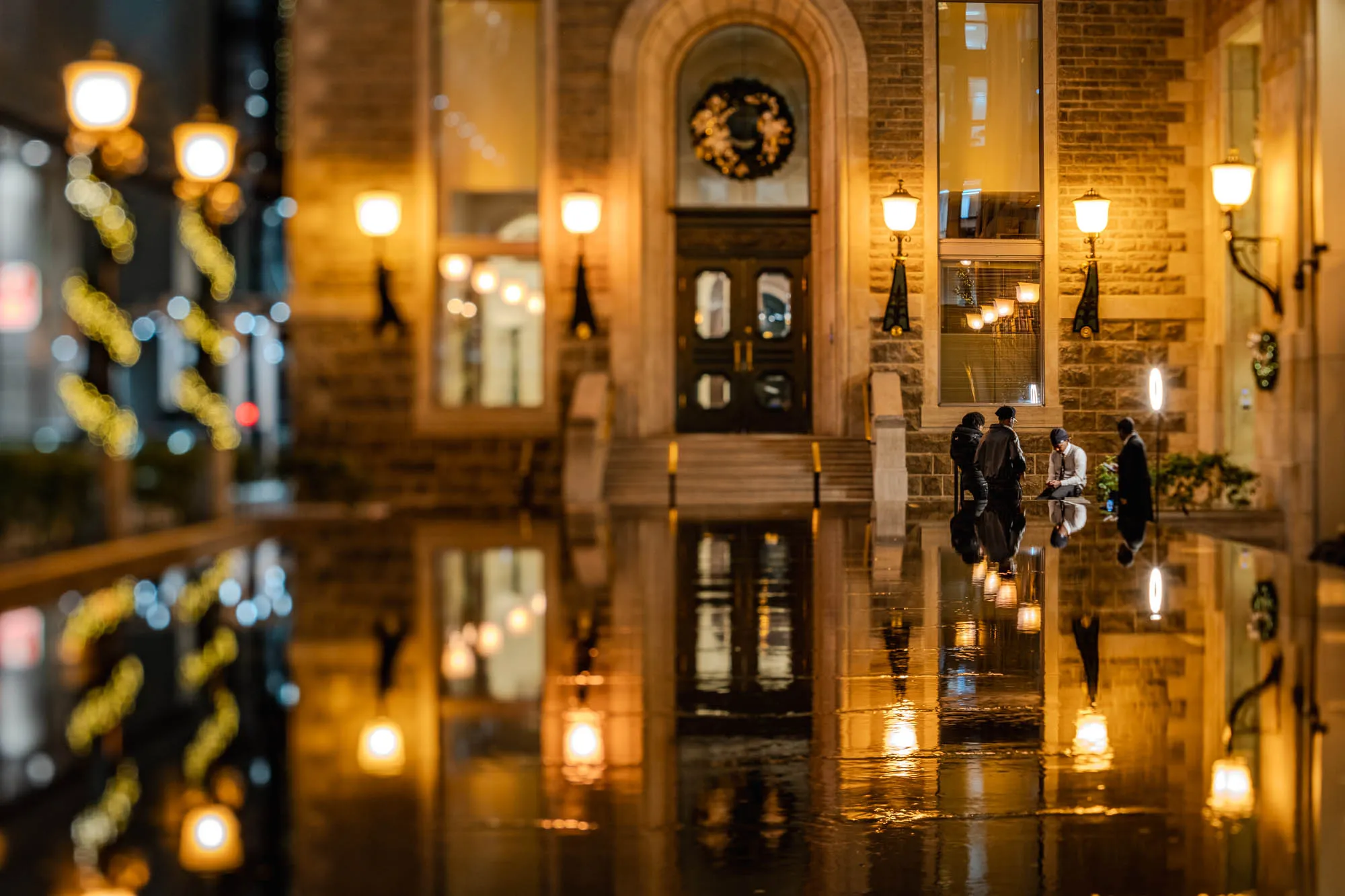 Four individuals next to a glistening reflecting pool at night, perfectly reflected below a stately stone building. Three figures stand, while one person kneels, focused on something at ground level, illuminated by a tall, glowing vertical ring light to their right. The building's arched entryway, adorned with a dark wreath, and its warm interior lights are mirrored in the slick ground, alongside several ornate wall sconces casting a golden glow. A very shallow depth of field renders distant street lights and decorative string lights as soft, ethereal orbs, contributing to the intimate nocturnal atmosphere.