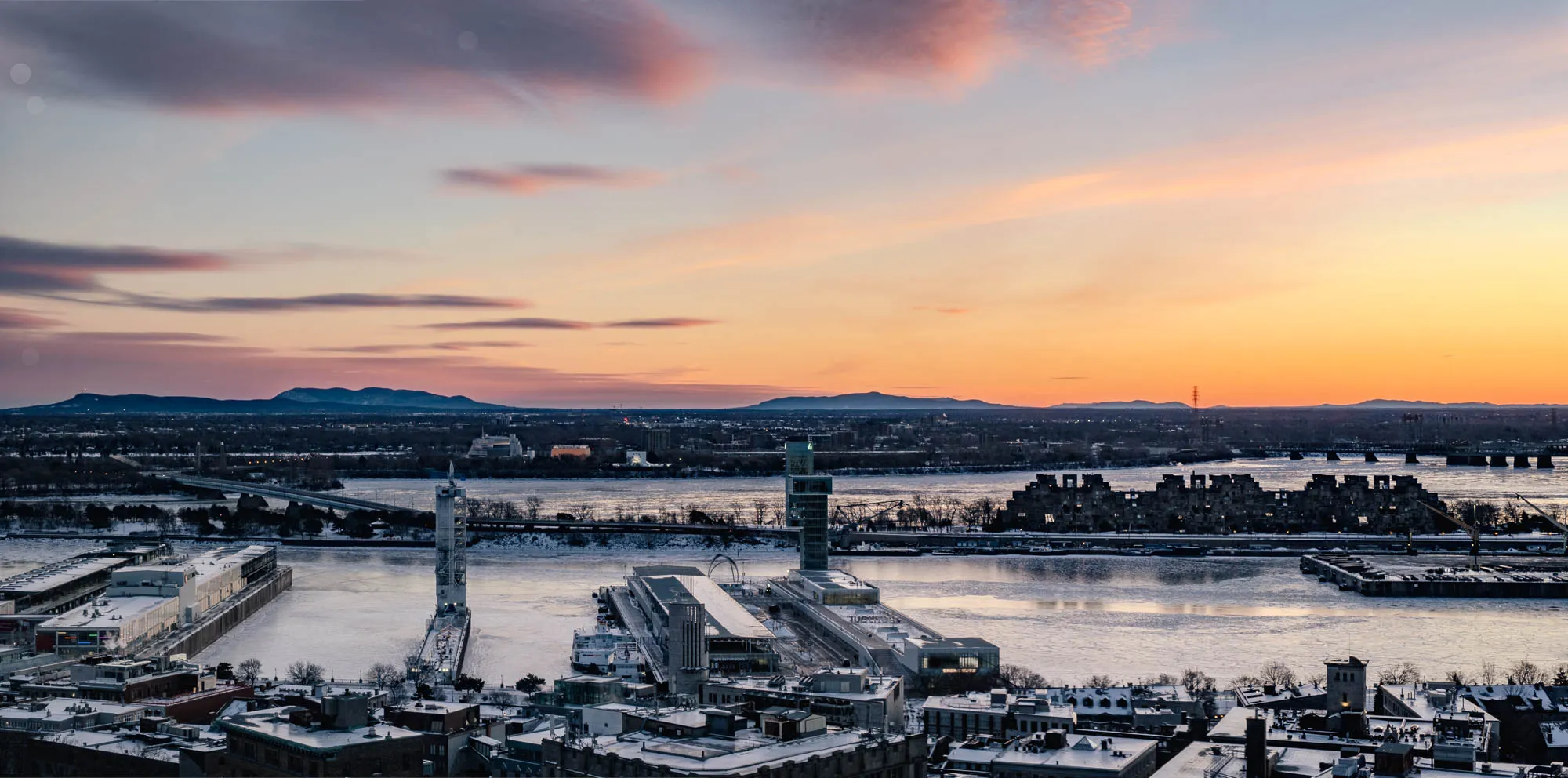 An expansive aerial view captures a frozen river cityscape at dawn, bathed in the soft, transitioning hues of a winter sky. In the foreground, snow-dusted rooftops line the riverbank, giving way to a wide, ice-covered waterway reflecting the faint light. Distinct modern structures punctuate the scene, including two tall observation towers on the near bank and a prominent cubist residential complex across the river, all sharply rendered by the deep depth of field. Distant, snow-capped mountains and a bridge span the horizon beneath a sky shifting from pastel pink and purple on the left to vibrant orange and yellow on the right.