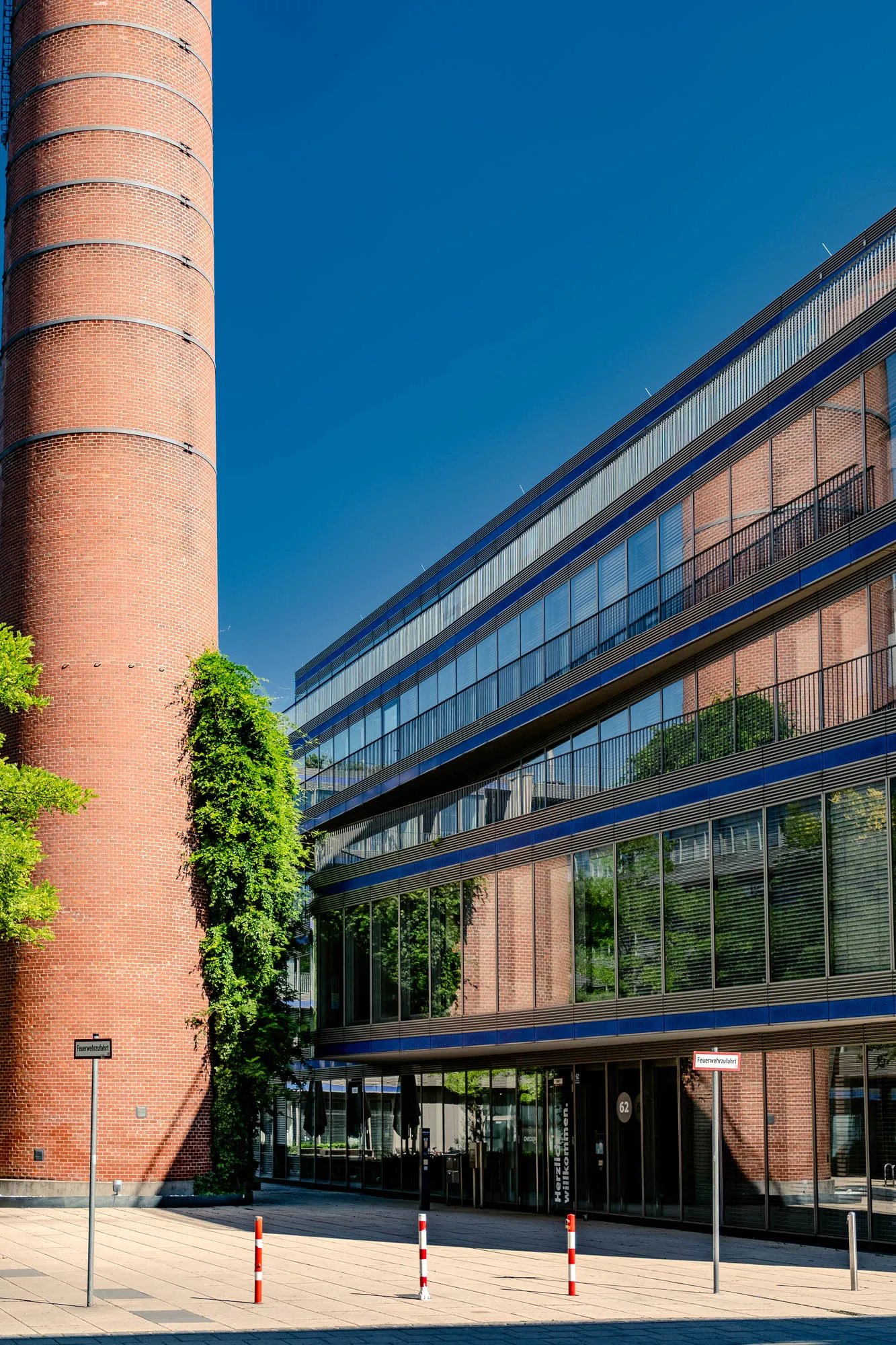 The image shows a scene with contrasting architectural elements under a clear blue sky. A tall, cylindrical brick chimney dominates the left side of the frame. The chimney has visible metal bands encircling it at regular intervals and green foliage climbing up part of its surface, partially obscuring the brick. To the right of the chimney, a modern, multi-story building stands with a facade primarily composed of reflective windows and visible brick in the background. The building's horizontal lines are emphasized by blue accents running along each story, along with metal accents. The windows reflect the chimney and other trees, blending the old and the new. The ground is paved with light-colored stone tiles, and several bollards with red and white stripes are present. Signage is visible near the building's entrance and on the left side near the chimney, though the text is difficult to read.