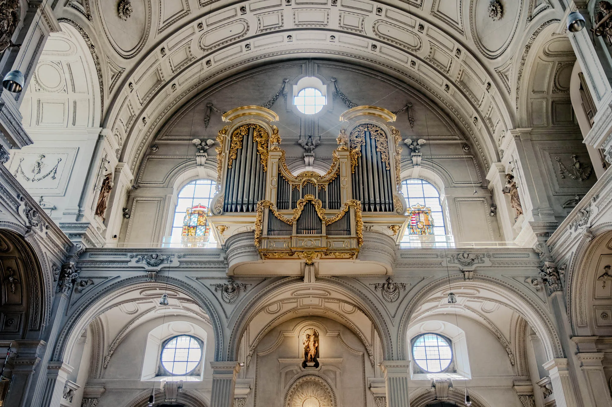 The image shows the interior of a grand church, looking upward towards a magnificent pipe organ. The church's walls are a pale gray, intricately decorated with moldings, sculptures, and arches. Sunlight streams through several arched windows, illuminating the details. Above, the vaulted ceiling is ornate, with recessed panels and decorative plasterwork. The pipe organ, centrally positioned on a balcony, is a focal point with its array of vertical pipes and ornate gold detailing. Statues are positioned along the walls at different points, adding to the sacral ambiance. The overall impression is one of architectural grandeur and spiritual significance.
