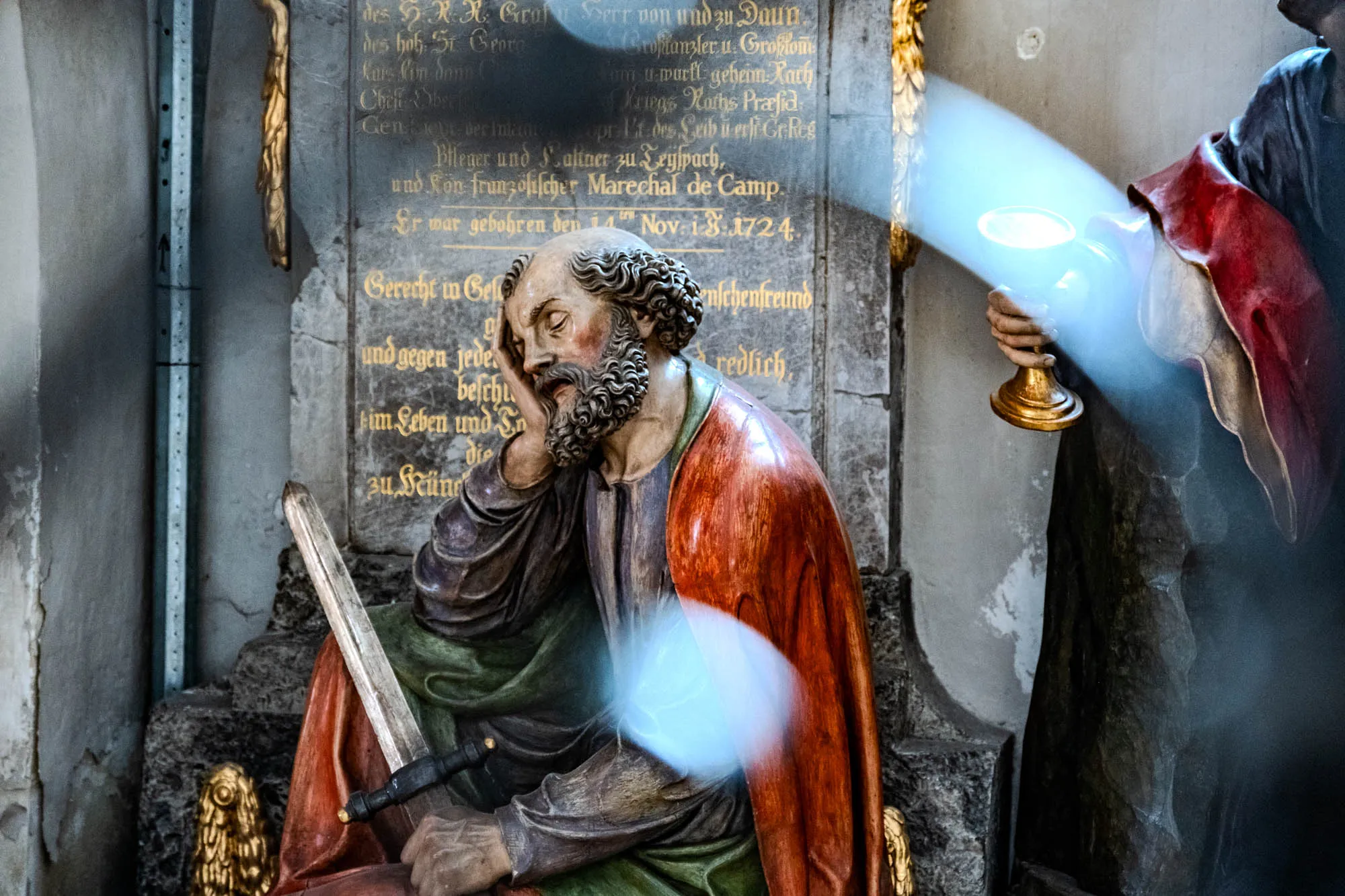 The photograph shows a detailed stone sculpture, likely part of a tomb or memorial, with intricate details. The sculpture depicts a man, possibly a soldier or saint, seated with his head resting on his hand in a contemplative pose. He has a full beard and curly hair. His clothing is draped over his body, featuring a red and orange cloak. A sword rests beside him on the stone surface. The sculpture is set against a stone background, which appears to be an inscribed memorial tablet. Text in German or a similar language is etched into the stone. To the right of the seated figure, another figure is partially visible, holding a golden object, possibly a chalice or lamp. The photograph's lighting creates a dramatic effect, with highlights emphasizing the textures of the stone and the figures' clothing. There are also some blurry patches due to either the camera settings or a natural phenomenon, casting light over the scene.