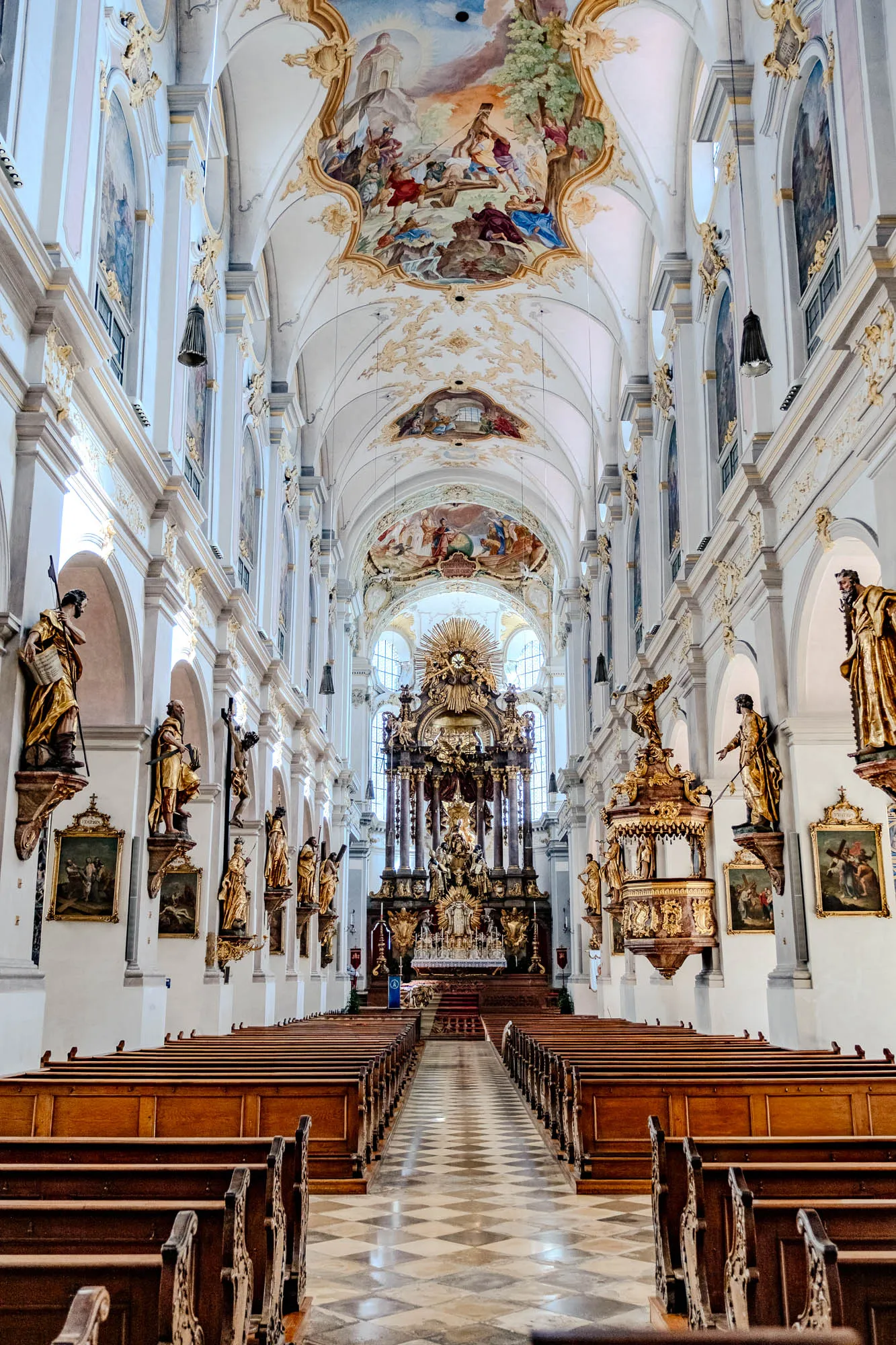 The image showcases the interior of a grand, ornate church, captured from the perspective of someone standing near the entrance, looking towards the altar. Rows of dark wooden pews fill the center aisle, leading the eye toward the back. The floor is covered in a light-colored checkered pattern. Flanking the pews on both sides are white walls with arched alcoves housing golden statues of figures, each mounted above framed paintings. Elaborate gold filigree adorns the walls. Above, the ceiling is a masterpiece of art, featuring colorful frescoes depicting religious scenes. The ceiling arches high, creating a sense of vastness and grandeur. At the far end of the church, the altar is a towering structure of gold, intricately carved and brightly lit, dominating the space.