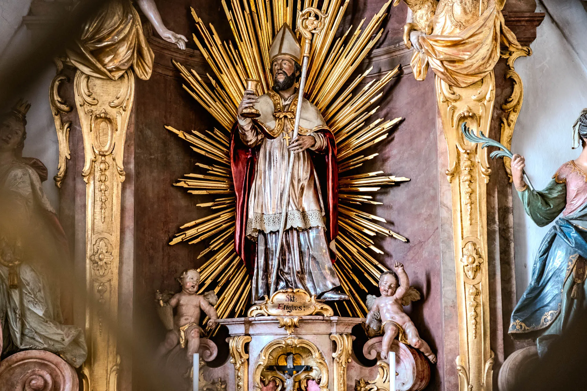 The photograph depicts a detailed, ornate religious altar. At the center stands a painted wooden sculpture of a bearded saint, likely Saint Eligius, identified by an inscription below the sculpture. He wears a tall, peaked hat, a flowing cloak with a red lining, and holds a golden chalice in his left hand and a long staff topped with an intricate design in his right hand. Behind the saint, radiating outwards, is a golden sunburst effect, comprising of many individual rays of light. The statue stands atop a decorated pedestal. Flanking the central figure on either side are cherubic figures, sculpted and painted with a pinkish hue. To the left and right, elaborate gold-painted architectural details frame the composition. On the far left and right, partial views of other sculptures are visible, draped with carved cloth, adding to the sense of depth and opulence. The overall tone is one of reverence, history, and artistic craftsmanship.