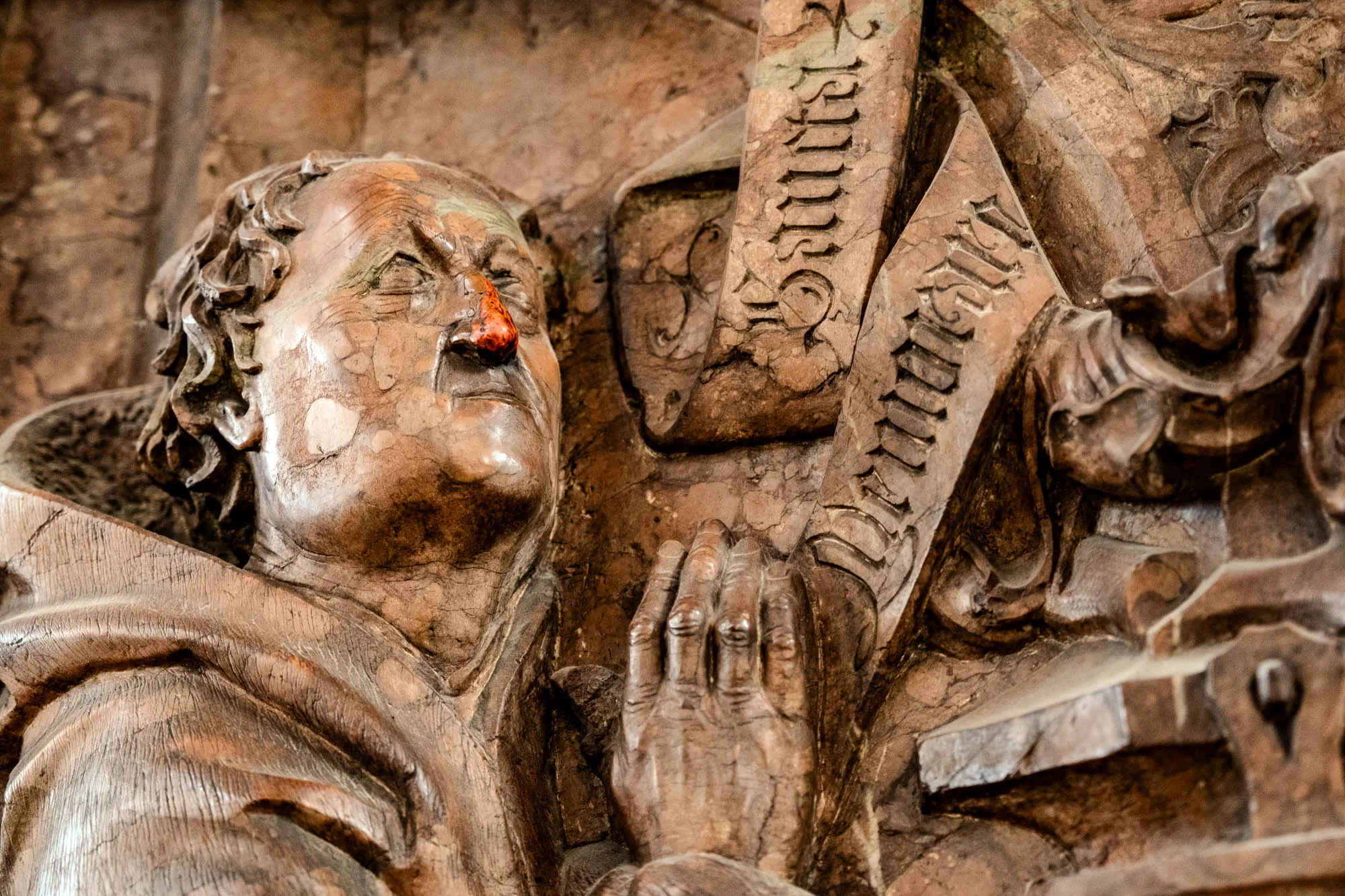 This is a detailed close-up of a carved wooden sculpture of a man, likely part of a larger religious or historical scene. The man has a slightly furrowed brow and looks upwards. He's wearing a cowl-necked robe, suggesting a religious figure or monk. The wood grain is prominent, giving a sense of age and texture to the carving. To his right, a carved hand is raised in what could be a gesture of supplication or prayer, with intricately detailed fingers. The background consists of other carved elements, including what appears to be a banner or scroll with inscribed text. The text is illegible, but the decorative style suggests an old script. The overall impression is one of somber reflection and intricate craftsmanship, emphasizing the fine details of the sculpture.