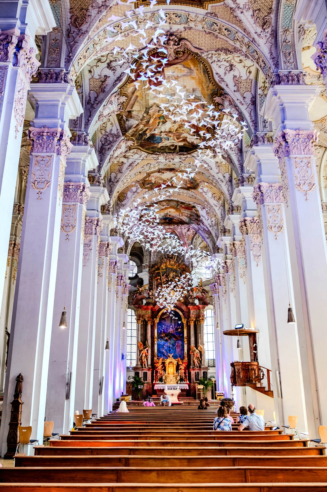 The image showcases the interior of a grand church, viewed from the back. Rows of tiered wooden benches fill the foreground, leading towards the altar. Several people are seated on the benches. Flanking the central aisle are towering white pillars, intricately adorned with ornate carvings and moldings. Above, the vaulted ceiling is a masterpiece of artistry, featuring detailed frescoes painted in muted earth tones. Suspended from the ceiling are two large installations resembling flocks of birds, possibly made of white paper or a similar lightweight material. These bird-like structures add a whimsical and modern touch to the otherwise classical and baroque setting. The altar is elaborately decorated with gold leaf and colorful sculptures, bathed in soft light from a large window behind it. A dark wood pulpit sits to the right, contrasting with the bright columns and ceiling.