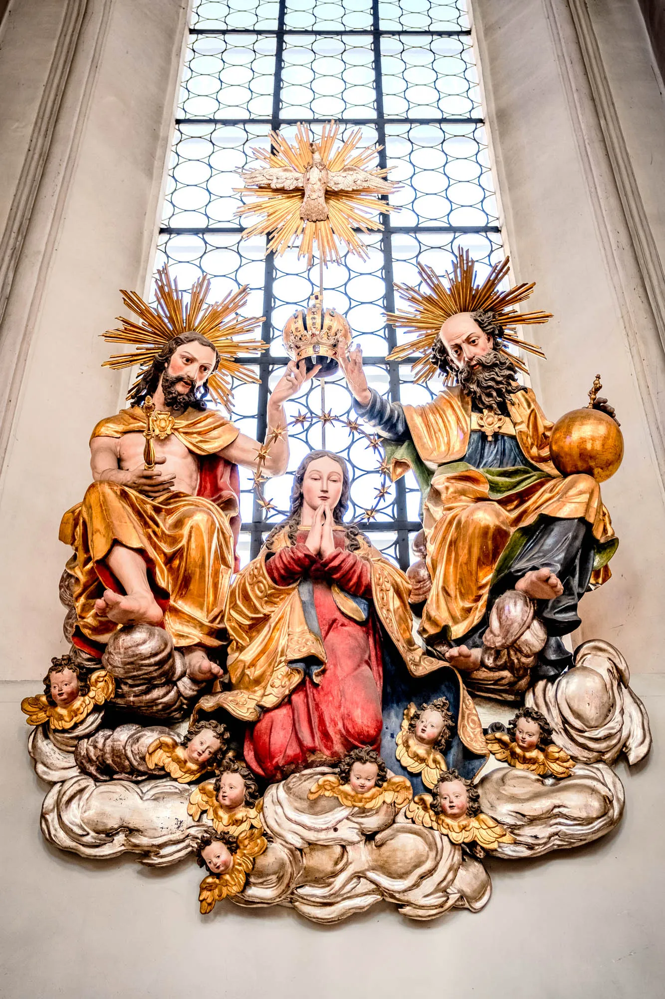 A detailed, ornately carved and painted religious sculpture is positioned in front of a large window. The sculpture depicts the Coronation of Mary by the Holy Trinity. Two male figures, representing God the Father and Jesus Christ, sit on either side of Mary, who is kneeling with her hands in prayer. They are all seated atop fluffy, silver-white clouds populated by multiple cherubic angels with golden wings. God the Father, on the right, has a long beard and holds a golden orb, symbolizing dominion. Jesus, on the left, is younger and holds a golden cross. Both figures wear draping, golden cloaks and have halos of golden rays emanating from behind their heads. Above the group, directly in front of the window, a golden dove with outstretched wings symbolizes the Holy Spirit. The window behind them has a lattice-like pattern. The sculpture's colors are rich and varied, from the reds and blues of Mary's robes to the golden hues of the other figures' attire and halos. The sculpture appears to be made of wood and is meticulously detailed, creating a sense of depth and texture. The background is the soft grey of the church wall and the brightness from the window. The sculpture and window are positioned high up on the wall.