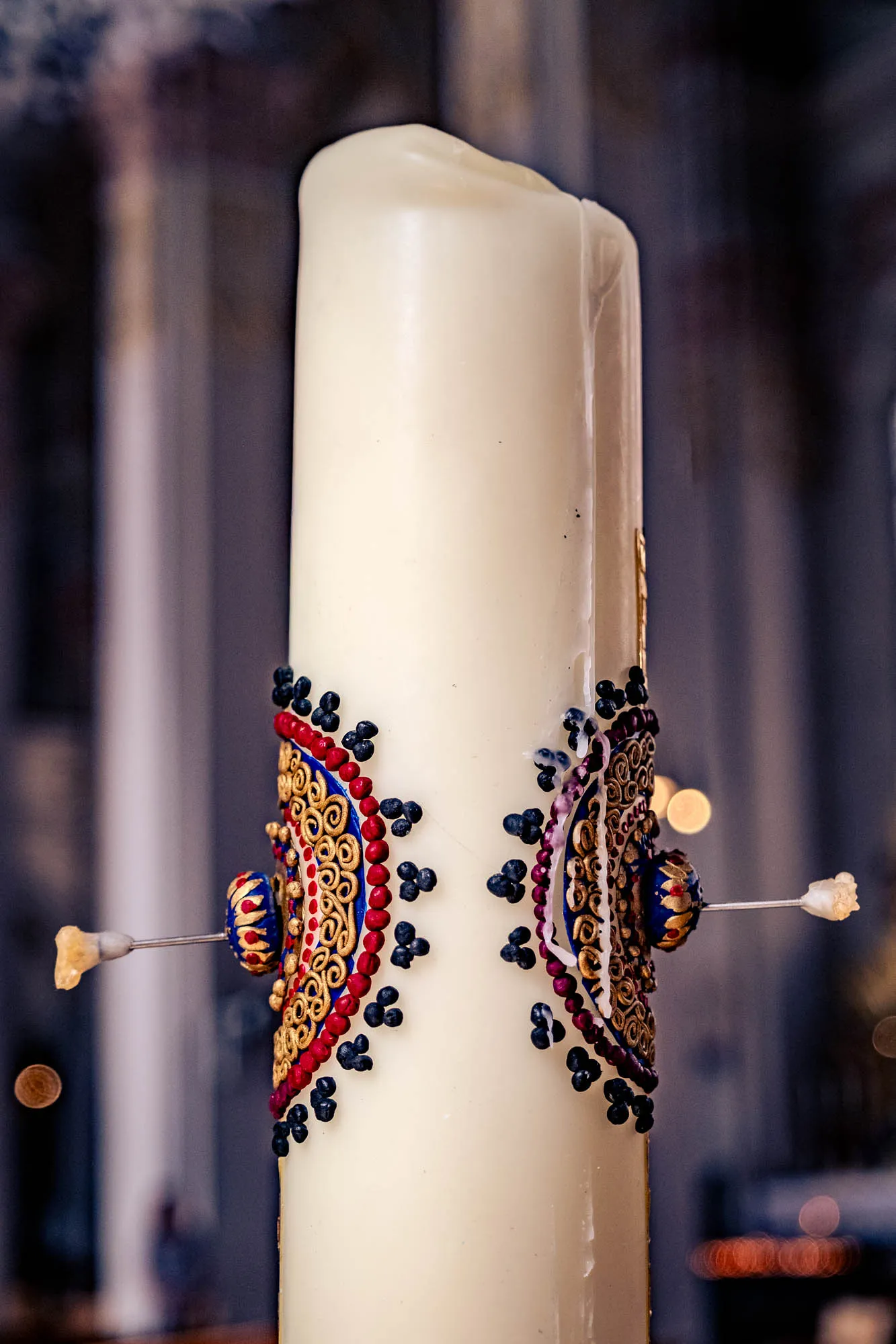 A close-up of a large, ornately decorated white candle. The candle is cylindrical and unscented, its surface smooth and slightly glossy. Around its midsection, there is a decorative band with two identical half-circular emblems. These emblems are highly detailed, featuring a gold filigree pattern bordered by lines of small red beads. These red beads are also accented with tiny clusters of three small, dark purple beads that seem to drip down the candle. The emblems are attached to the candle with a pin that is visible on the left and right side and end with a small dark blue and gold bead. The pin shows a bit of wax clinging on the left side of the candle. The candle and decoration sit against a soft-focus backdrop of what appears to be an architectural interior, perhaps a church or cathedral, with vertical lines suggesting columns and blurred light points creating a sense of depth.