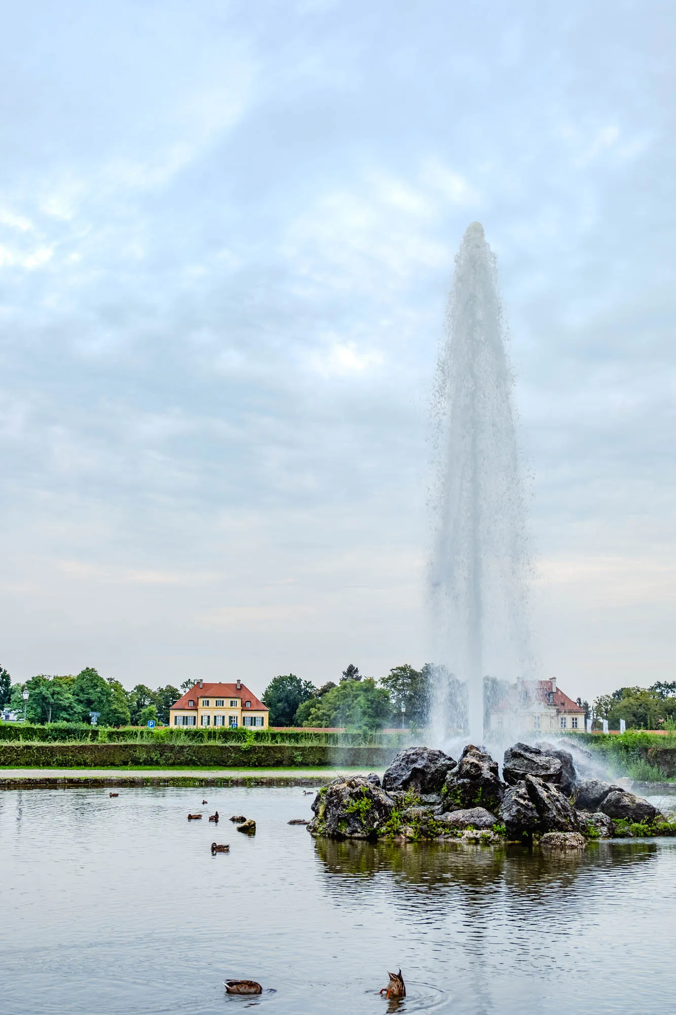 The image captures a serene scene of a large fountain in the middle of a calm lake, framed by a soft, overcast sky. The fountain erupts forcefully, shooting a tall column of water high into the air. The base of the fountain appears to be a cluster of large, dark rocks, some covered with patches of green moss or small plants. The water falls back down around the rocks, creating a misty spray. Several ducks are visible on the lake, some swimming peacefully while others appear to be diving or feeding. In the background, across the lake, a yellow building with a red-tiled roof stands out against a backdrop of green trees. The building is framed by a manicured hedge and a few other buildings can be seen in the distance. The sky is filled with soft, gray clouds, suggesting a peaceful, perhaps slightly cool day.