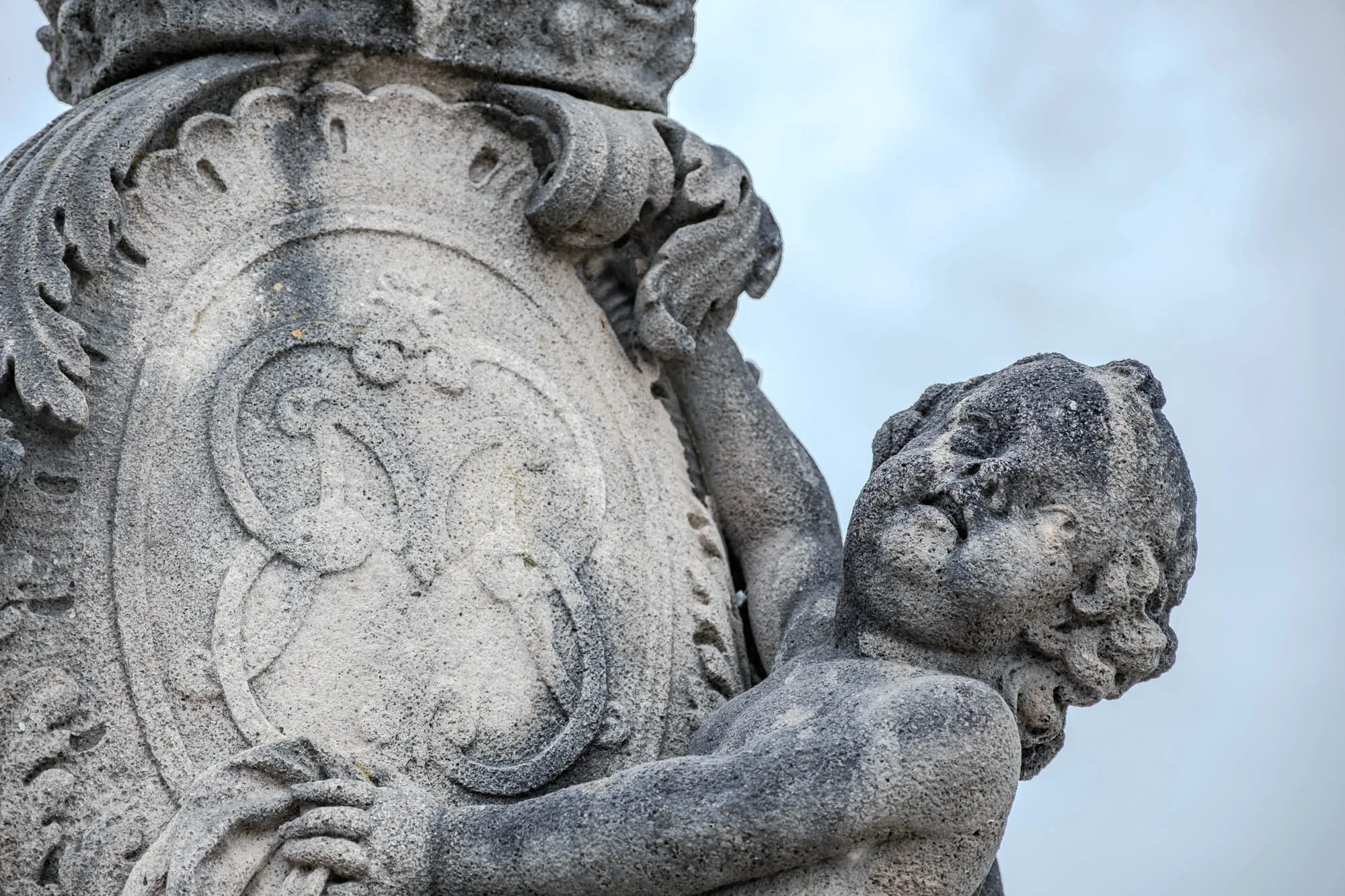 The image shows a close-up of an old stone statue, likely part of a larger architectural element. The statue features a cherubic figure clinging to a large, ornate shield or cartouche. The child figure, rendered in textured stone, has rounded features and appears to be supporting the shield with one arm wrapped around it and the other hand grasping its side. The shield is heavily decorated with raised patterns, including what appears to be floral or leafy designs around the edges, and a more abstract pattern within the center circle, which has a raised circle to further distinguish the design. The stone surface shows signs of age and weathering, with visible texture and discoloration, lending to the overall antique appearance. The background is a soft, blurry sky. The lighting accentuates the three-dimensional form of the sculpture.