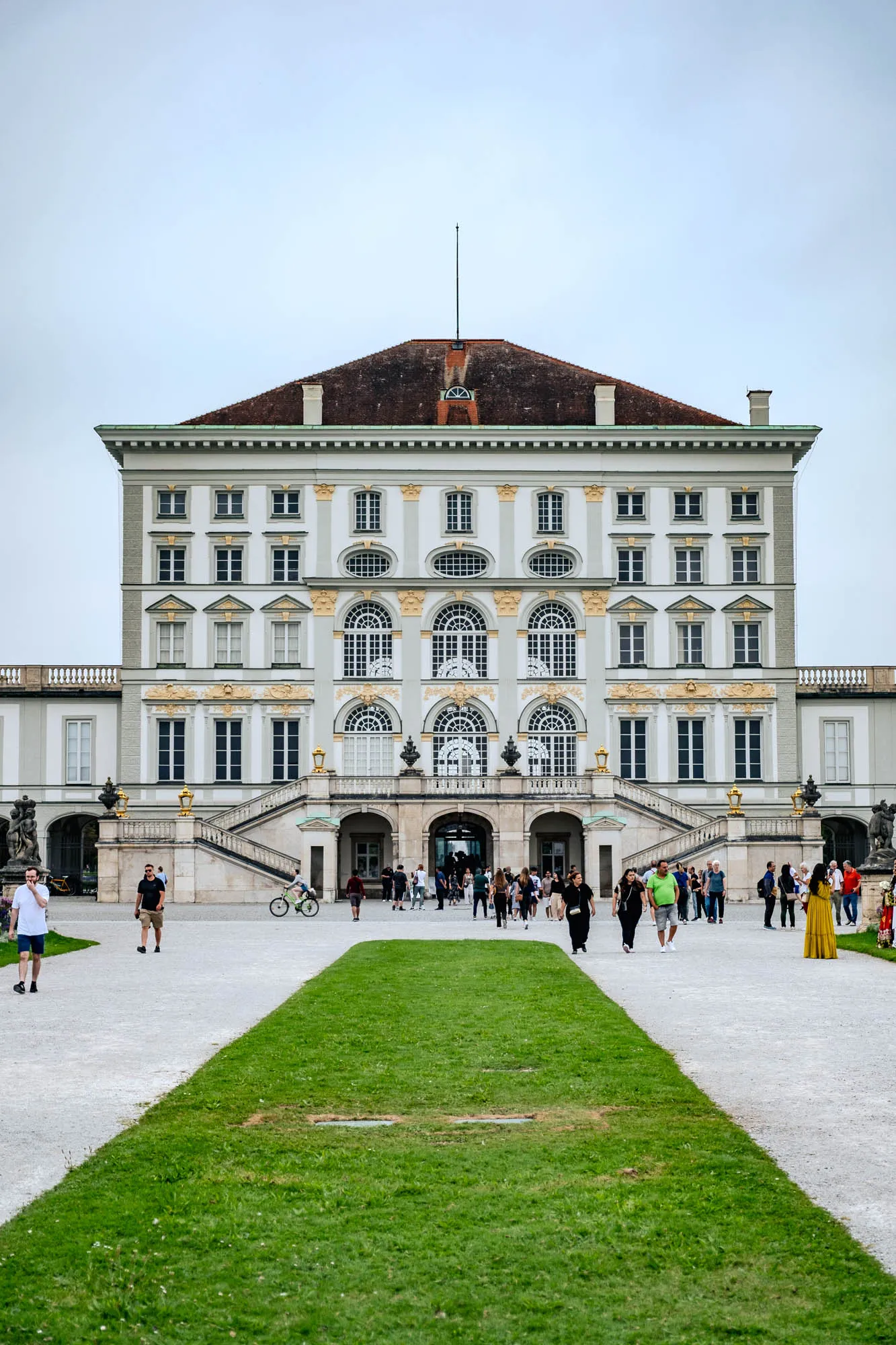 The image captures a grand, symmetrical view of Nymphenburg Palace, a large, light-colored baroque building. The palace's facade features multiple stories of windows, some rectangular and others arched, with decorative golden accents around the arches. Two symmetrical staircases sweep up to the palace entrance from either side. A red-tiled roof with a small spire sits atop the building. Below the palace, a long, green lawn stretches towards the viewer, dividing the gray stone courtyard. People stroll across the courtyard in both directions, adding a sense of scale and life to the scene. The sky is overcast, creating a soft, diffused light.