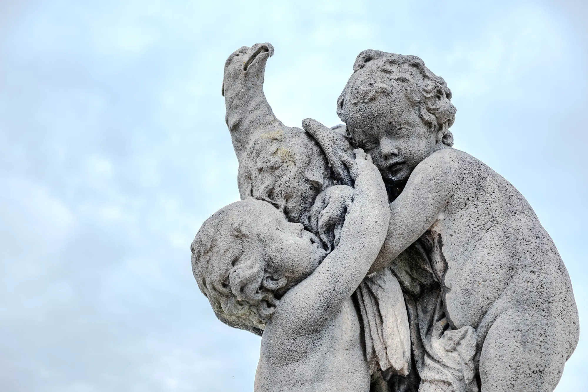 A weathered stone statue depicting two young cherubs and a goose stands against a soft, diffused sky. The cherub on the right is slightly larger, with plump features and curly hair, embracing the goose tightly. Its face is turned slightly downward, its expression pensive. The second, smaller cherub is nestled close, its face partially obscured by the goose's neck, creating a sense of protectiveness or perhaps a shared secret. The goose, with its long neck arched gracefully, appears alert and watchful. The statue's surface is textured and aged, showing the subtle signs of exposure to the elements, suggesting its presence in an outdoor setting. The light sky provides a gentle backdrop, enhancing the statue's form and lending a timeless quality to the scene.