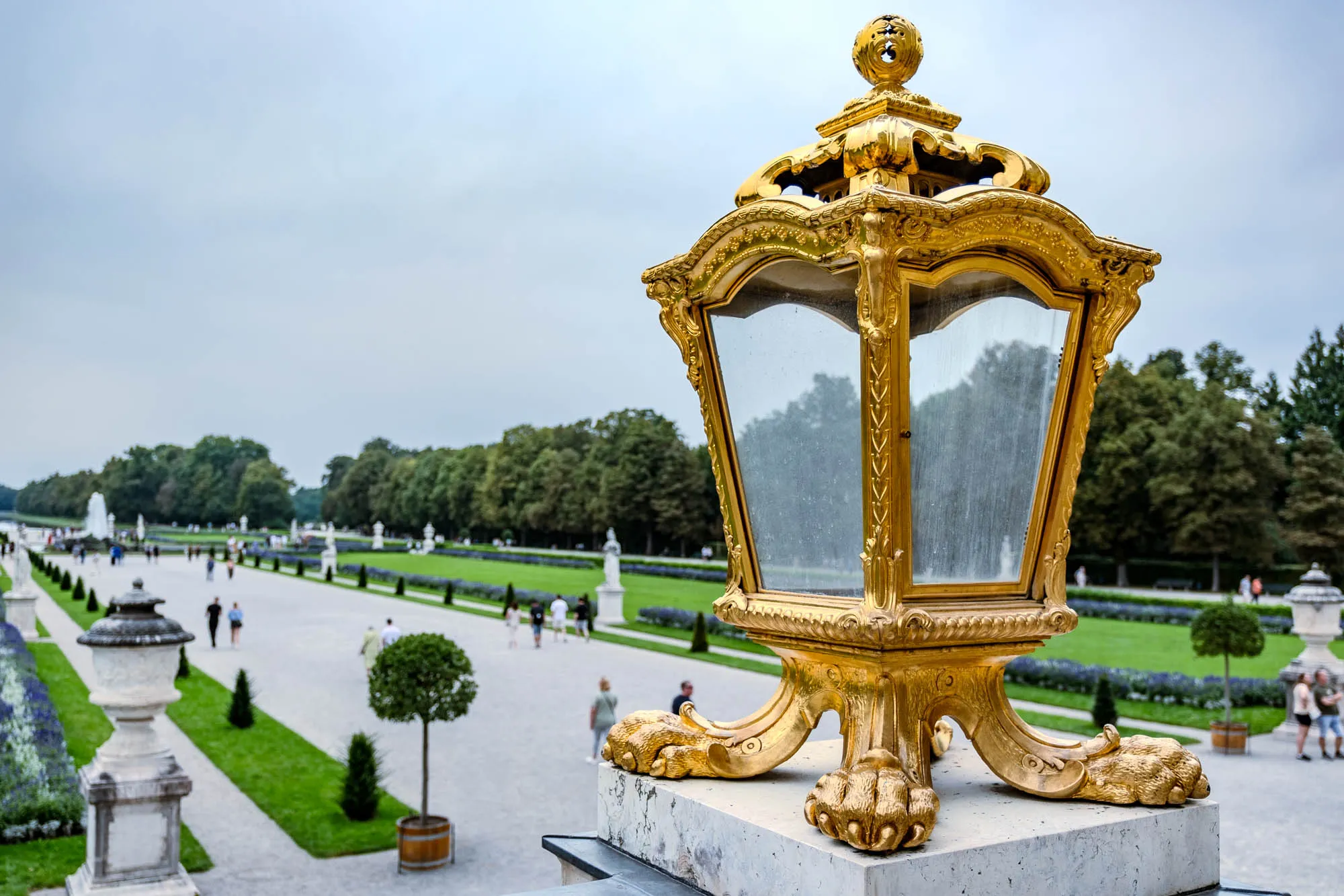 Close-up of an ornate, golden lamppost standing on a stone base. The lamppost has a rectangular shape with glass panels on each side. Its golden frame is intricately decorated with scrolling patterns and topped with a spherical ornament. The base of the lamppost features four stylized claw feet. In the background, there's a wide view of a manicured garden stretching into the distance. A large pathway extends through the garden, lined with rows of neatly trimmed trees and flowerbeds. People can be seen strolling along the path. Beyond the garden, a line of trees forms the horizon under a cloudy sky.
