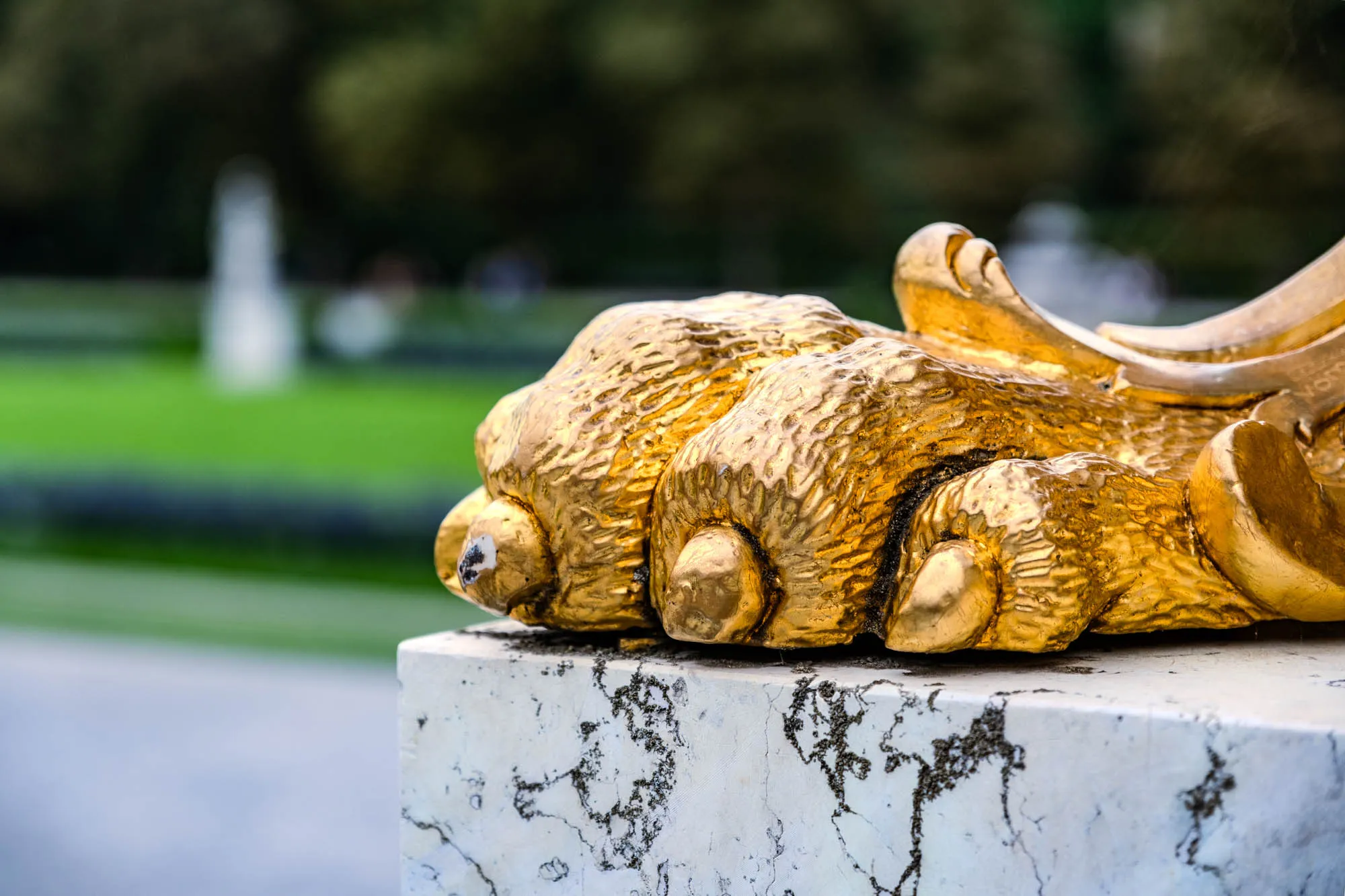 A close-up of a gilded, ornate sculpture rests on a weathered stone base. The sculpture features a realistic representation of a lion's paw, complete with defined toes and textured fur, all rendered in a bright, reflective gold. The paw is positioned as if it's firmly gripping the edge of the stone. The stone base exhibits a mottled, aged appearance with visible cracks and dark-colored lichen or stains, contrasting with the smooth, glossy gold of the sculpture. Behind the sculpture, the background is a blur of green foliage, suggesting a park or garden setting. The lighting highlights the three-dimensional details of both the sculpture and the stone, enhancing the contrast between the intricate artistry of the gold and the raw, natural texture of the stone.