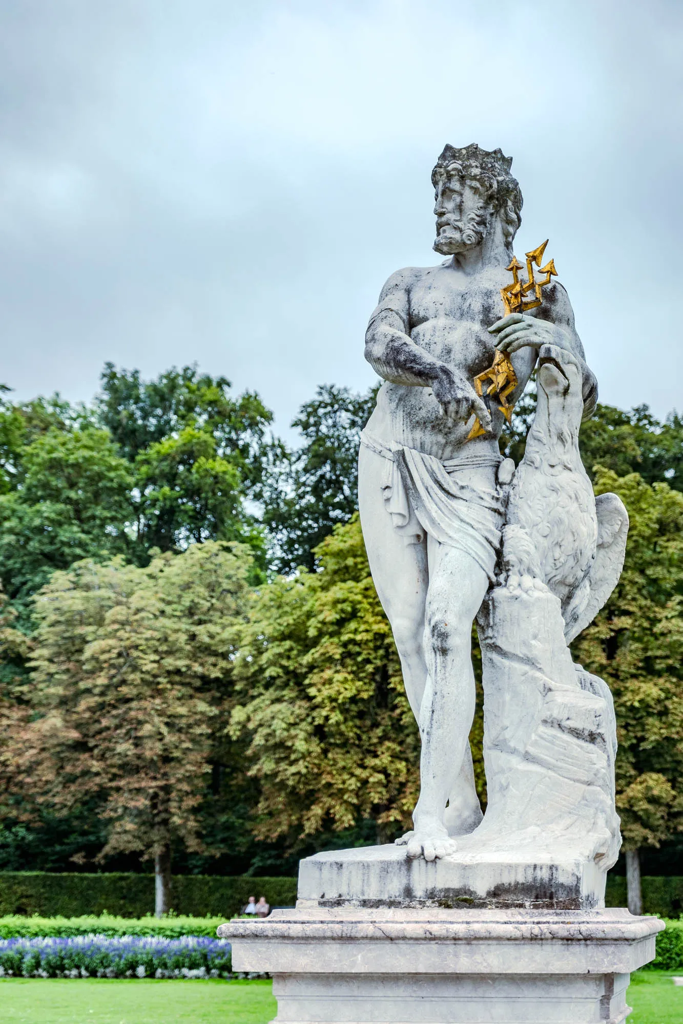 A full-length, stone sculpture of a classical figure stands against a backdrop of trees and cloudy sky. The figure, likely Neptune or Poseidon, has a weathered, greyish-white appearance, showing signs of age. He has a full, flowing beard and a crown of what appears to be seaweed or leaves on his head. His physique is muscular and draped with a cloth that wraps around his waist and partially covers his legs. In his left arm he holds a golden trident, and his right arm is resting upon a large, sculpted eagle. The eagle is also made of stone, with detailed feathers and a proud posture, its head reaching upwards towards the trident. The sculpture rests on a multi-tiered stone base, and the background features a lush, green garden with various trees and plants. The sky above is overcast, casting a soft, diffused light on the scene.