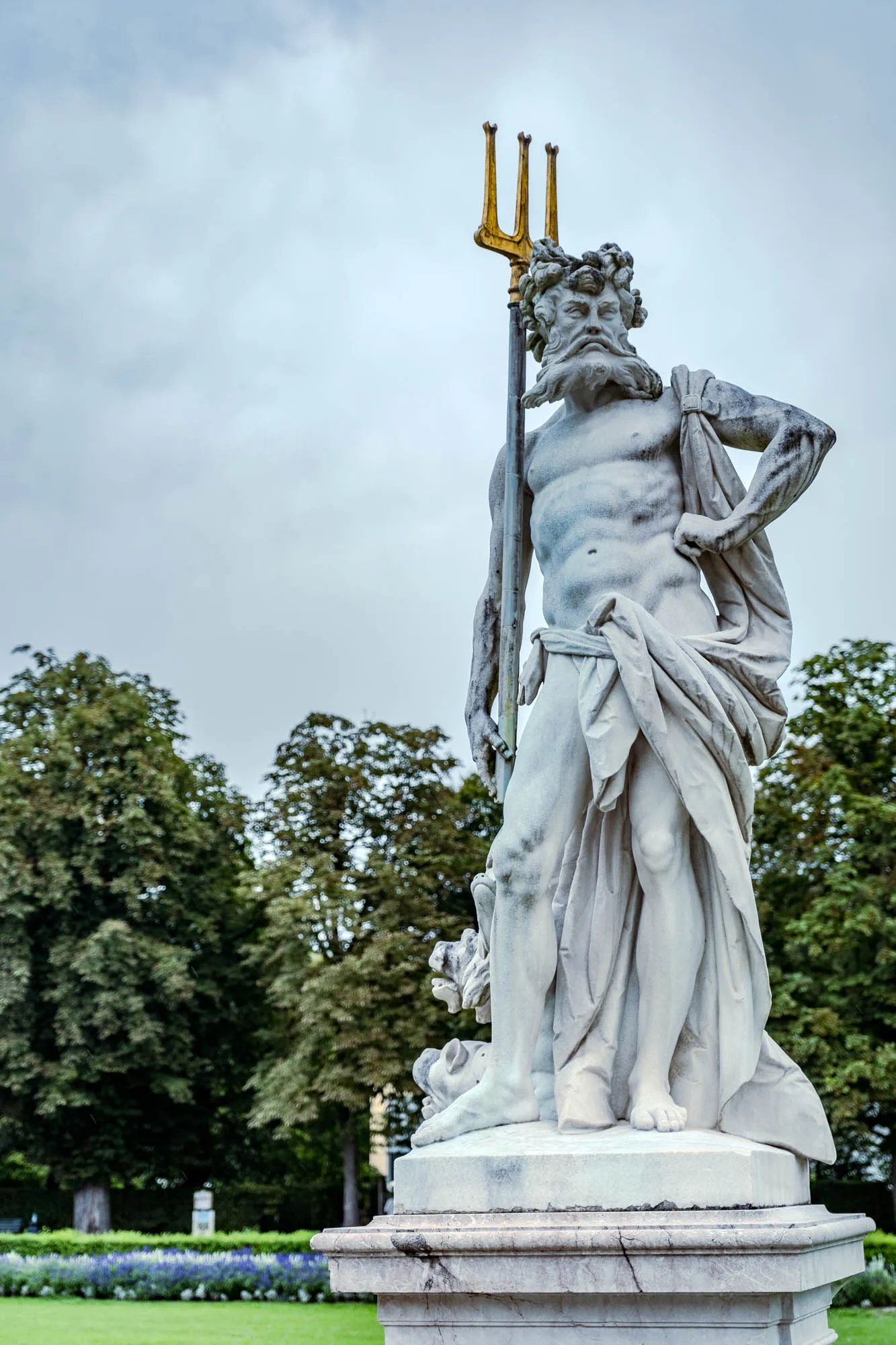 The image shows a full shot of a weathered, light-grey stone statue of a muscular, bearded man. He is standing upright on a tiered stone pedestal, his weight shifted slightly to his left leg. Draped across his right shoulder and waist is a piece of fabric, its folds and creases adding texture to the smooth stone of his body. In his left hand, he firmly holds a tall, golden trident, its three prongs pointing upwards. The man's facial expression is stern, his brow furrowed above deep-set eyes, and a long, flowing beard covers his chest. His hair is styled in tight curls, resembling seaweed or waves. In the background, there are lush green trees under a cloudy sky. At the bottom of the pedestal, there are detailed carvings of what appear to be sea creatures. The statue exudes strength and authority, a representation of a powerful figure, possibly a sea god.