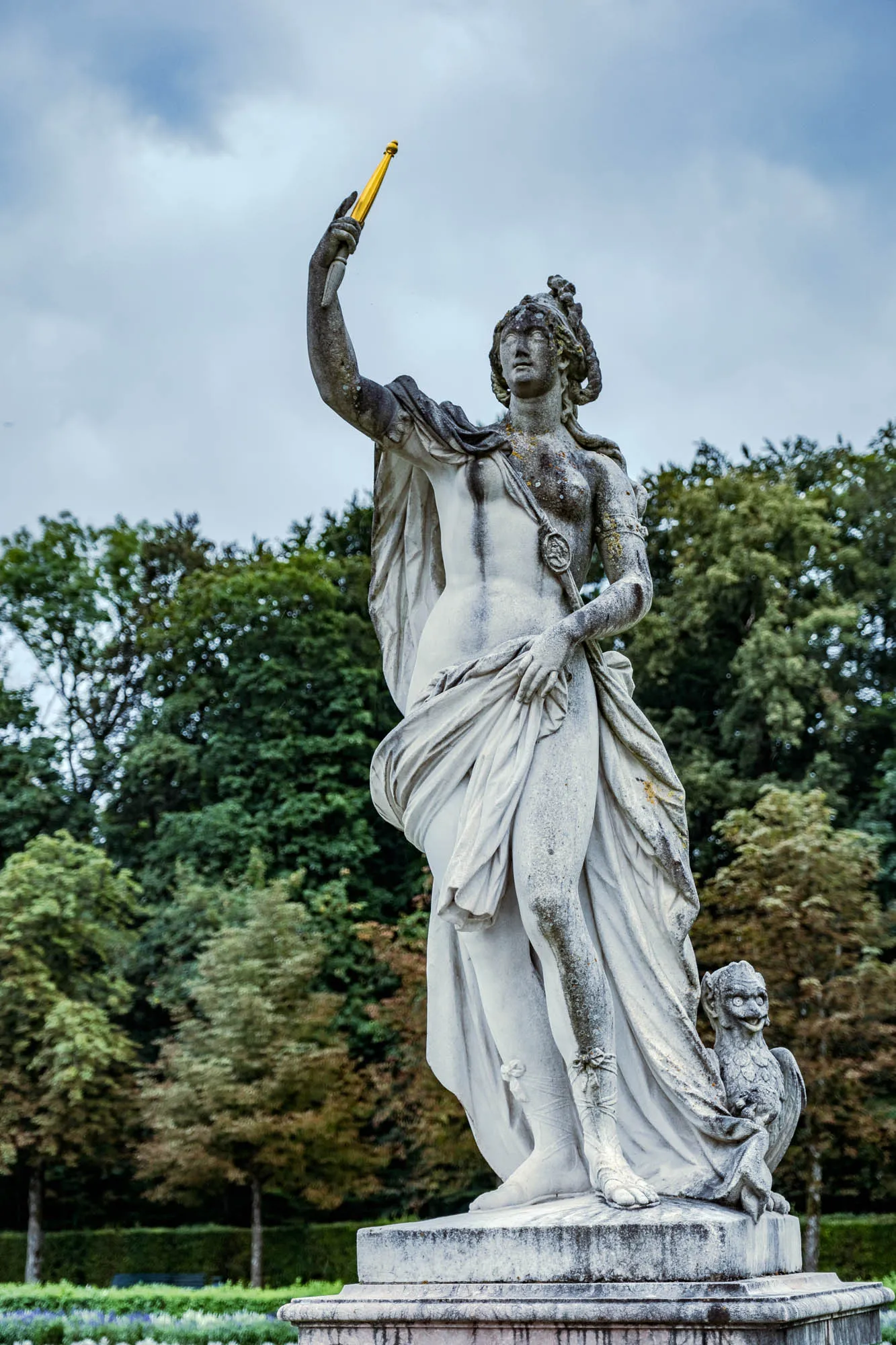 The image shows a full shot of a weathered stone sculpture of a female figure standing on a tiered stone base. Her body is draped in a flowing robe that clings to her form, revealing portions of her torso and legs. The robe is partially wrapped around her shoulders and falls down her back, giving the impression of motion. Her head is adorned with an elaborate hairstyle or headdress, and she holds a slender, golden object upright in her raised right hand, the object looking like a rod or wand. Her left arm is bent at the elbow and the hand rests gently near her waist. Next to her, on the right at base level, a smaller figure of a griffin is visible, its wings folded behind it. The statue appears to be old, with signs of age such as discoloration and wear visible on its surface. In the background is a scene of lush greenery, composed of many trees with varying shades of green foliage, suggesting a forest or park setting. The sky above is overcast with shades of gray and white.
