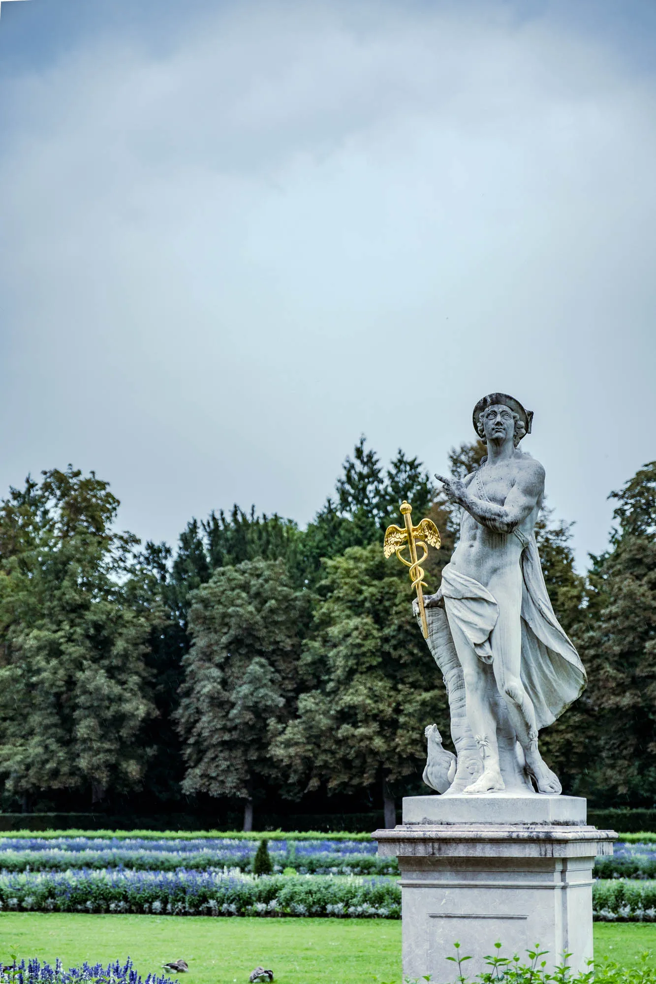 A full-length, eye-level shot depicts a weathered, light-gray statue of a classical figure, likely Mercury, standing atop a tiered gray stone pedestal. The statue is positioned against a backdrop of lush greenery and a cloudy sky. The figure is mostly nude, draped with fabric that gracefully falls from his left shoulder and across his legs. He wears a winged helmet or hat and holds a golden caduceus, the winged staff entwined with two snakes, in his left hand. His right hand is raised with the index and middle fingers extended. The statue appears aged, with visible signs of weathering. In the background, a vibrant green lawn stretches toward a thick line of trees. A row of low-lying, colorful flowering plants separates the lawn from the trees. The sky is overcast with gray clouds, providing soft, diffused light.