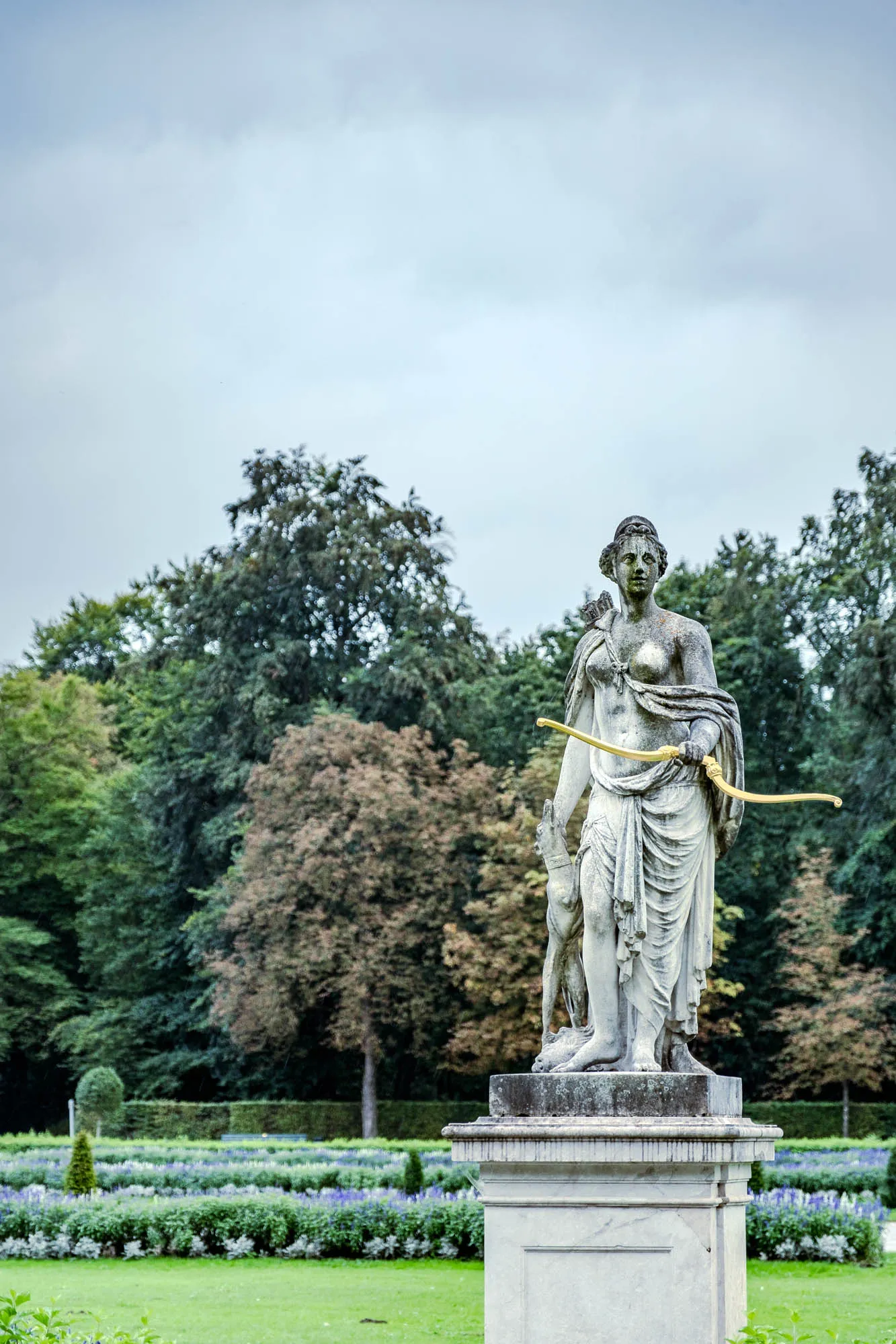 The image shows a full, medium shot of a classical sculpture of a female figure in a park setting. The sculpture appears to be made of a light-colored stone, possibly marble, and is weathered, showing signs of age and exposure. The figure is standing, draped in a robe that is gathered and flowing around her body, leaving her chest partially exposed. She holds a bow in her left hand, with the string visible. Behind her and to her right, a small deer stands at her side. The statue is mounted on a tiered stone pedestal. The backdrop features a well-manicured garden with neatly arranged rows of flowers and greenery. Beyond the garden, a dense forest of trees stretches into the distance under a slightly overcast sky. The overall impression is one of tranquility and classical beauty, with the sculpture serving as a focal point in the serene landscape.