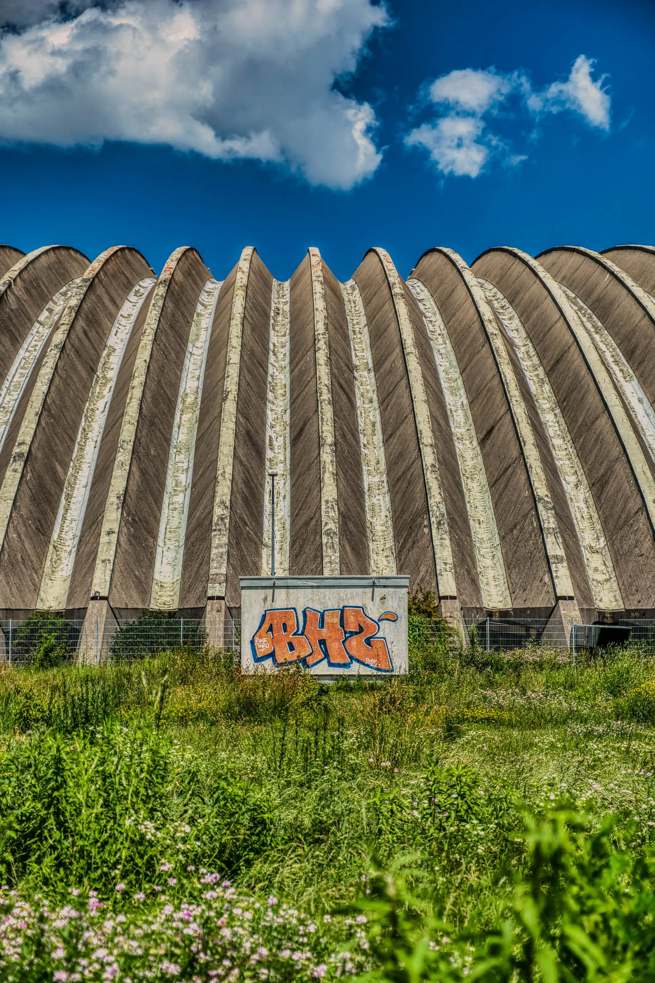 A concrete hanger-like arched warehouse with a rib-like facade, side-on, with a grassy meadow in front and a blue sky behind. A small square concrete building is in the center of the frame in front of the warehouse, with orange spray-painted letters on it