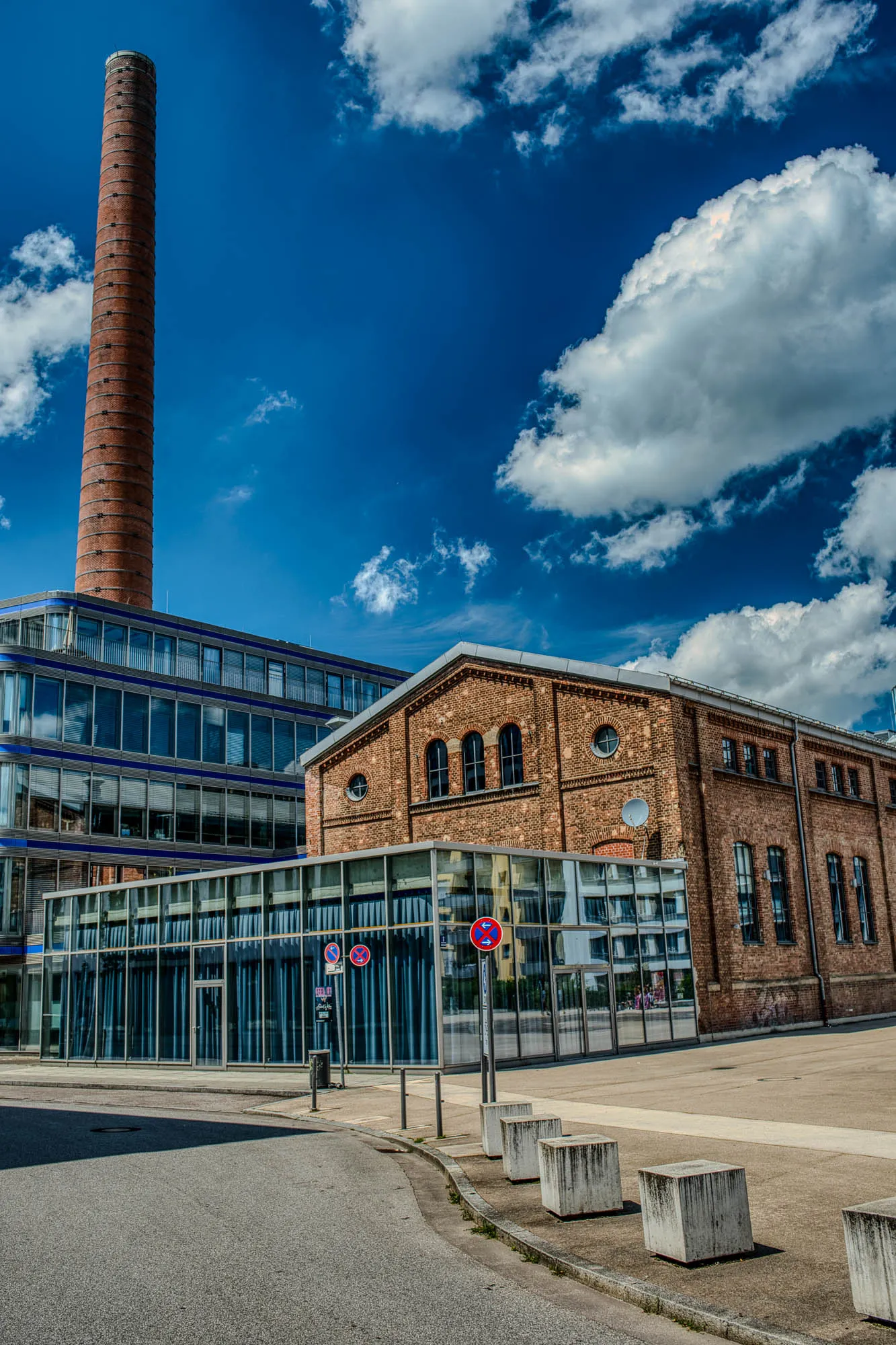 A single story glass building in front of a two story brick industrial building, with a five story glass building behind them, and an old smokestack behind the taller glass building. A street winds in front of them, with a plaza to the right