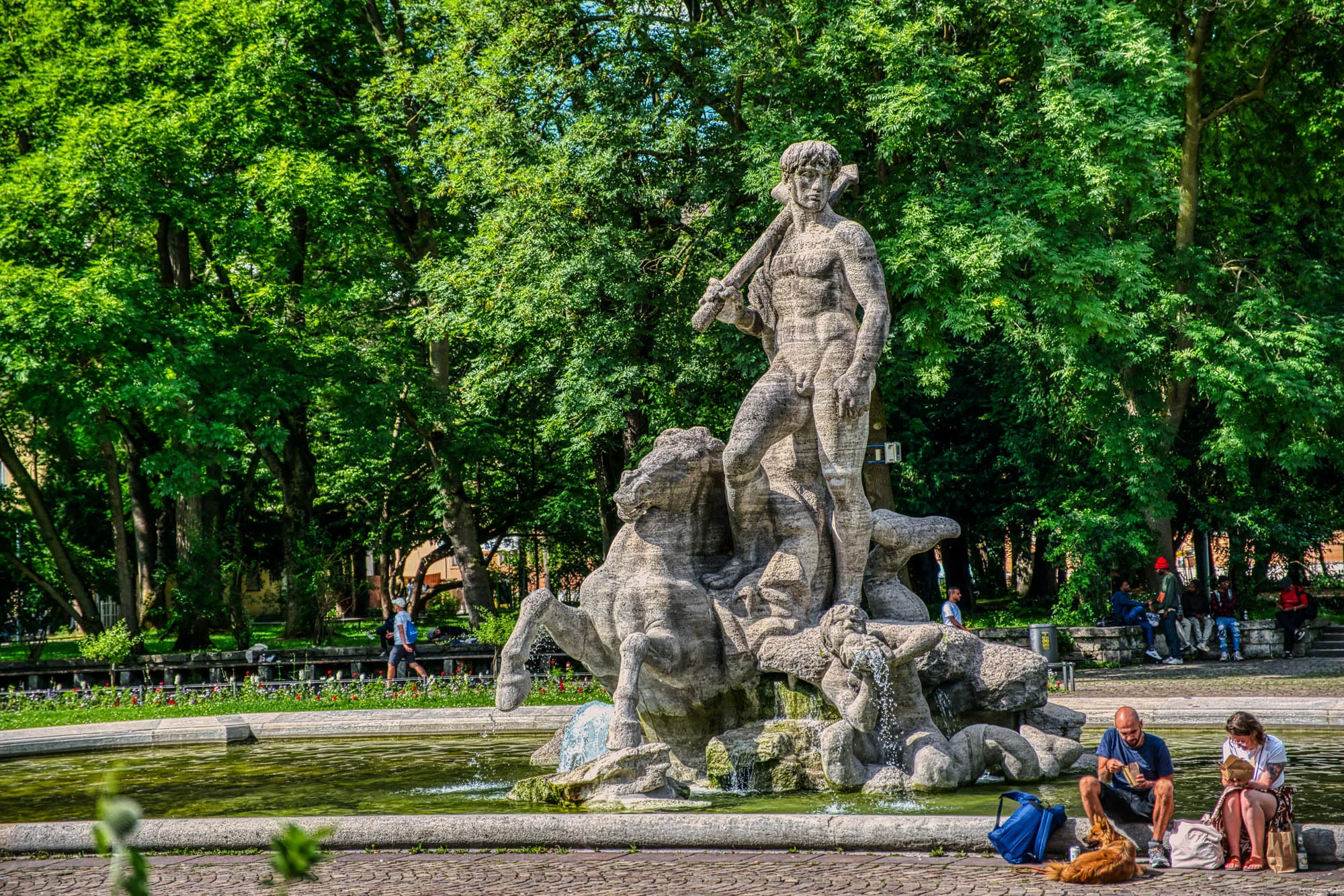 A stone fountain featuring Neptune as a nude man holding a trident on rocks, with a horse on his right, and a man crouching down blowing a shell that with water coming out, in the middle of a small pond. A couple and their dog sit on the lip of the fountain's pond, eating. Trees are in the background.