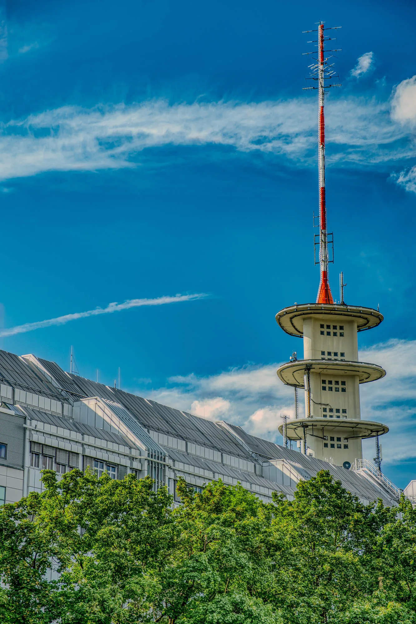 A radio tower, shaped like a triangle with three flat roof-like decorations spaced out up the side, peeking out from a roof line above the tree line. A red and white radio antenna sits above the tower.