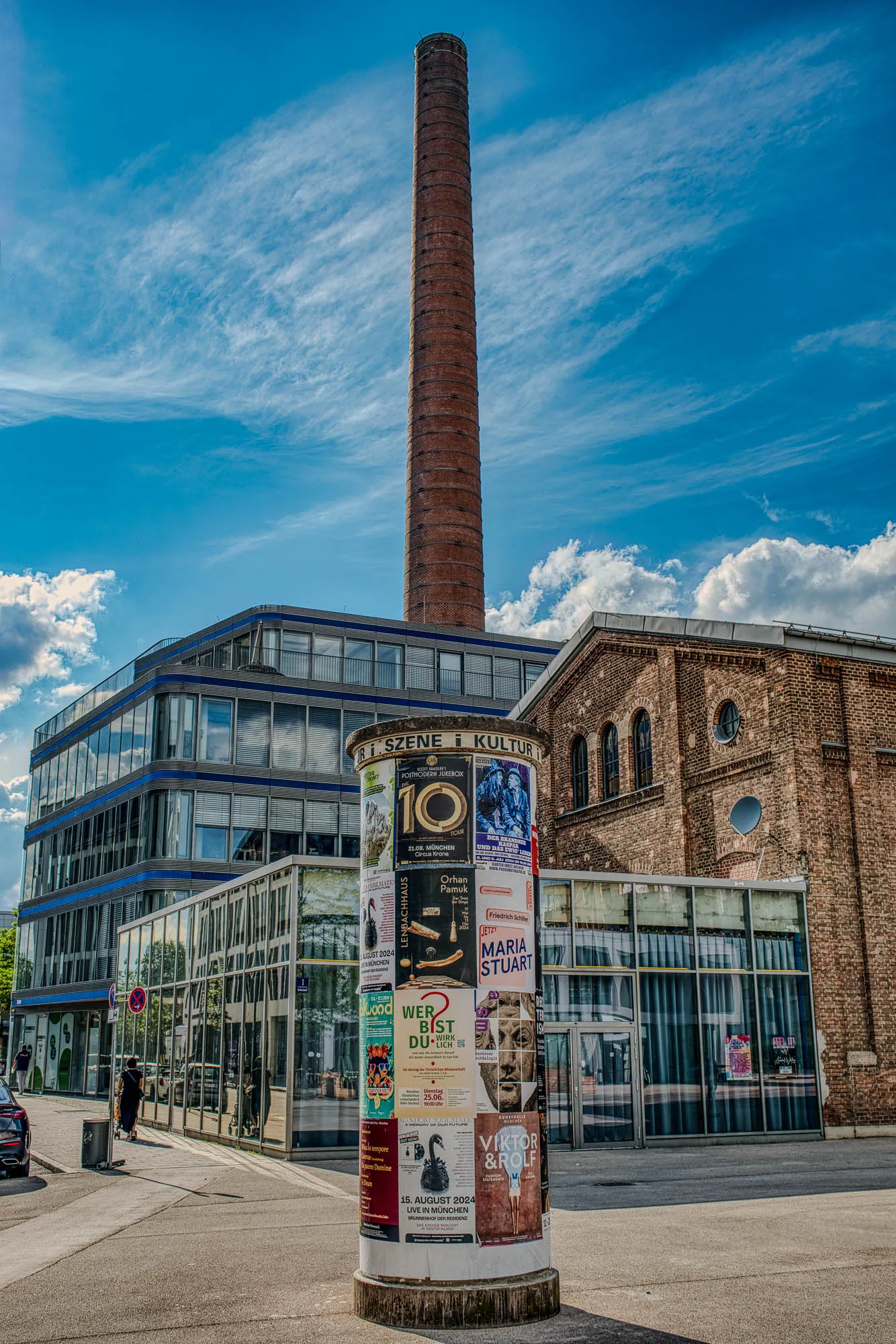 A public notice board, shaped as a cylindar, in front of a short glass building in front of a repurposed industrial brick building, in front of a new four story glass building, with an old industrial smokestack in the background. The notice board lines up with the chimney.