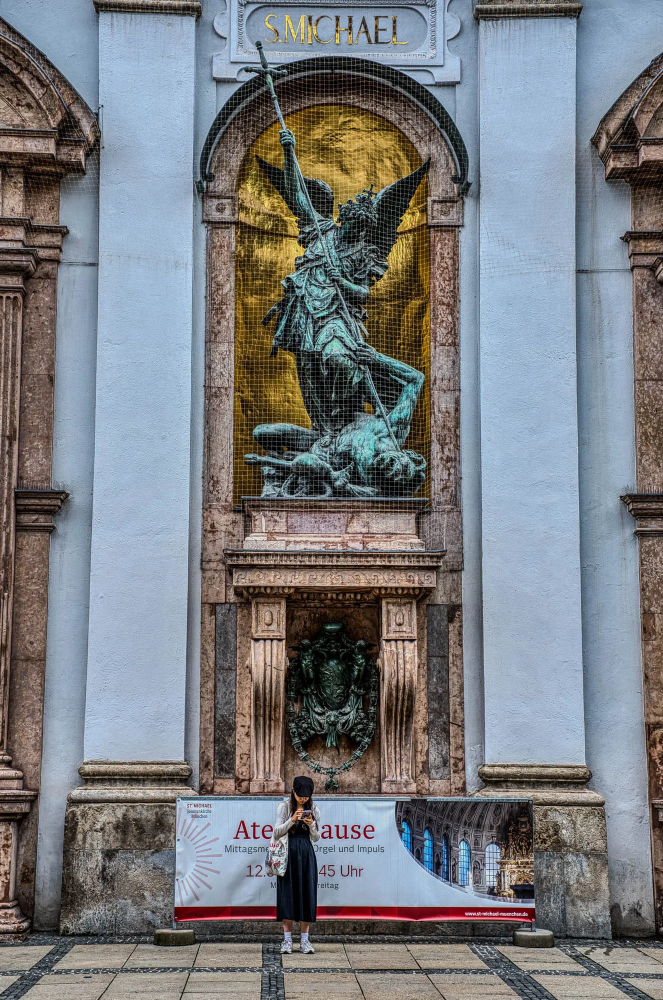 A patinated statue depicting St. Michael fighting a demon in front of a gilded carveout framed by marble, a woman standing underneath it looking at her phone. A patinated crest is underneat the statue.