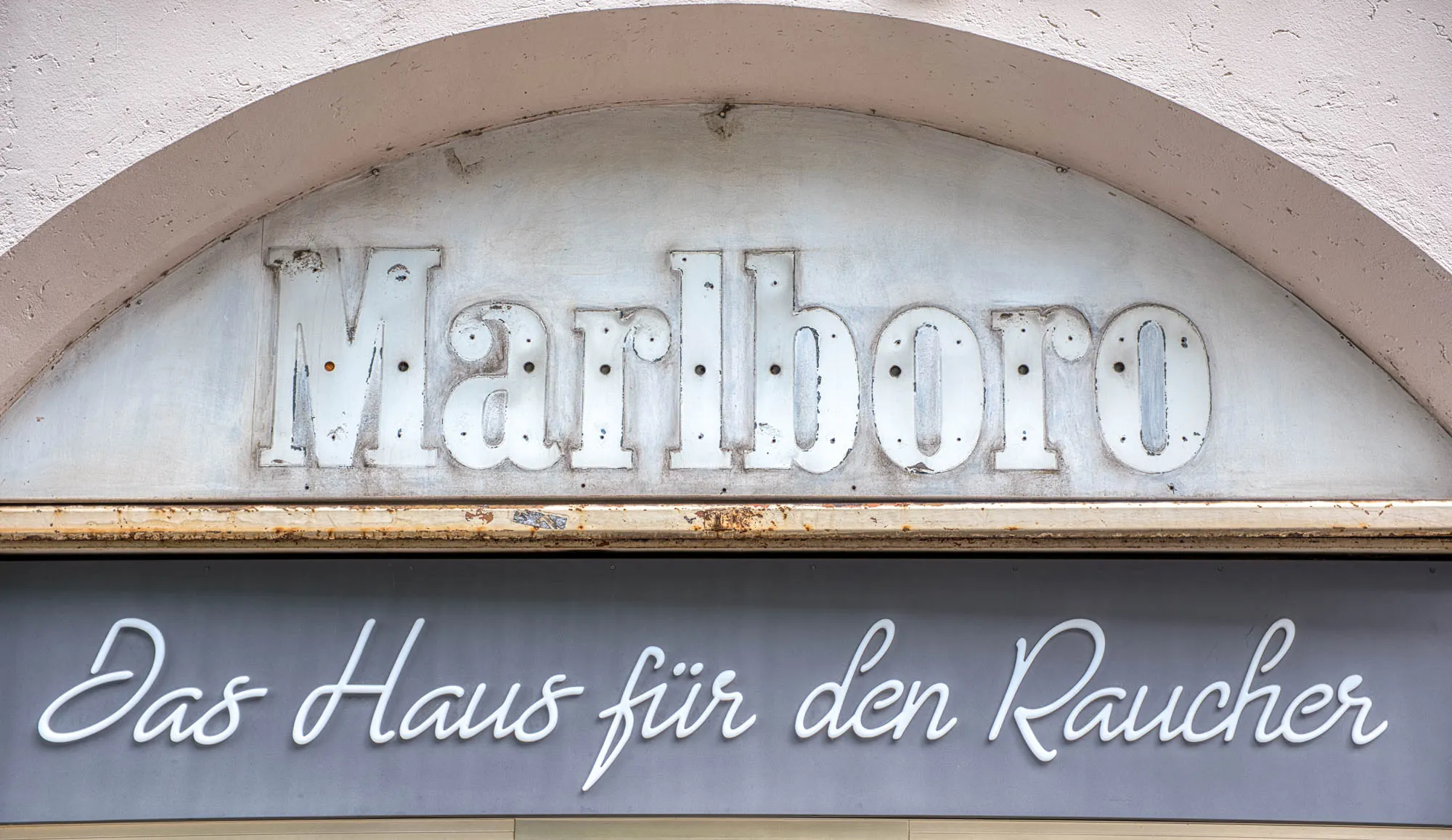 A closeup of a storefront in Munich that reads (in German) "the house for the smoker" in a white script. Above it, the silhouette of an old Marlboro sign that once hung.