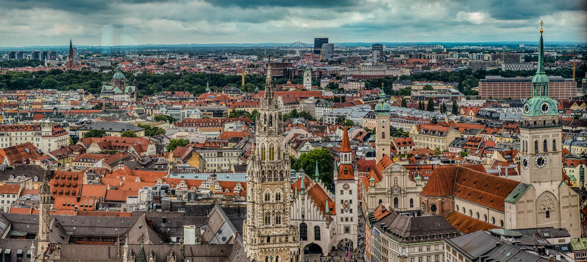 Sweeping panoram of old town Munich with the old town square in the foreground, a ferris wheel behind a line of trees in the mid ground, and hills in the far distance.