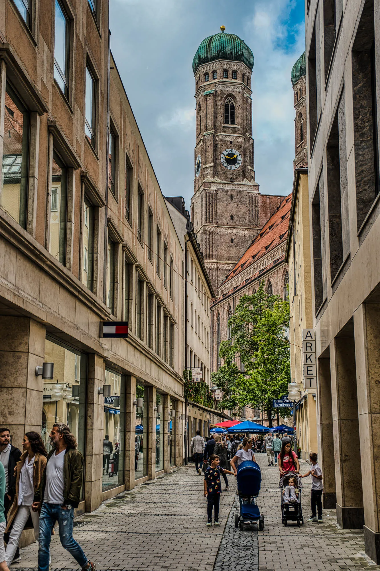 The blue and green dome-topped tower of Frauenkirche rising from the front of the church, a bustling alleyway framing it on either side, sight lines drawing you back towards the church and up to the tower.