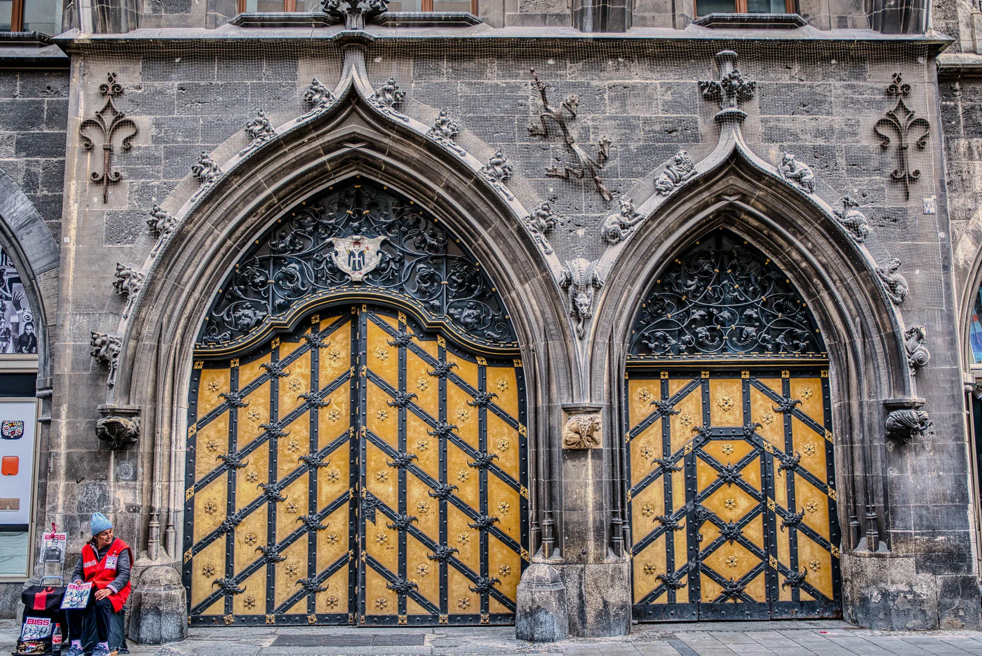 Two large archways with gilded doors on a stone building. A woman in a red vest sells BISS magazines in front of them.