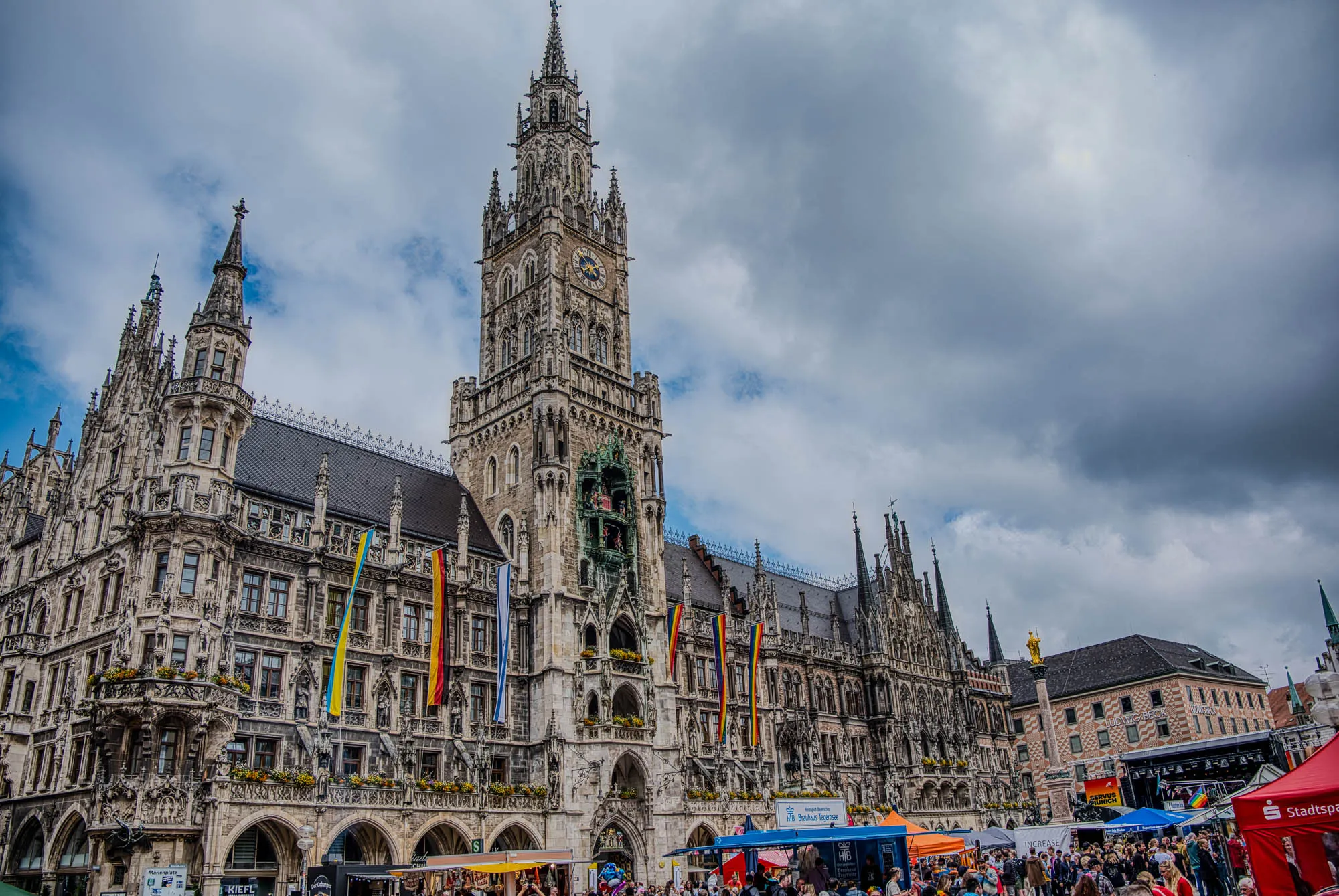 Neues Rathaus (new town hall) in Marienplatz, the historic town center of Munich. Its flying six flags, a Ukranian flag, a German flag, an Israeli flag, and three rainbow flags. In the center of town a Pride celebration and faire is happening.