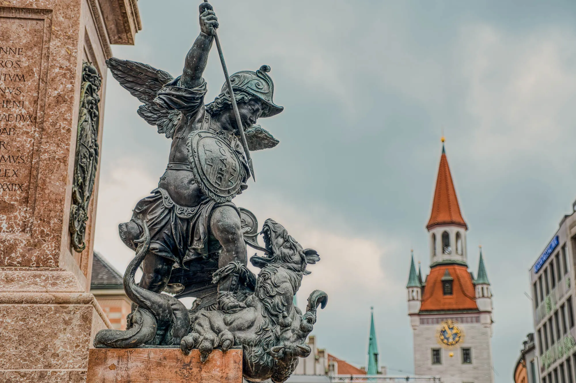 Small statue of a putto fighting a dragon depicted with the head of a beast, tallons, tentacles, and wings, on the marble podium base of the monument Mariensäule  in old town Munich. A clock tower is visible in the background