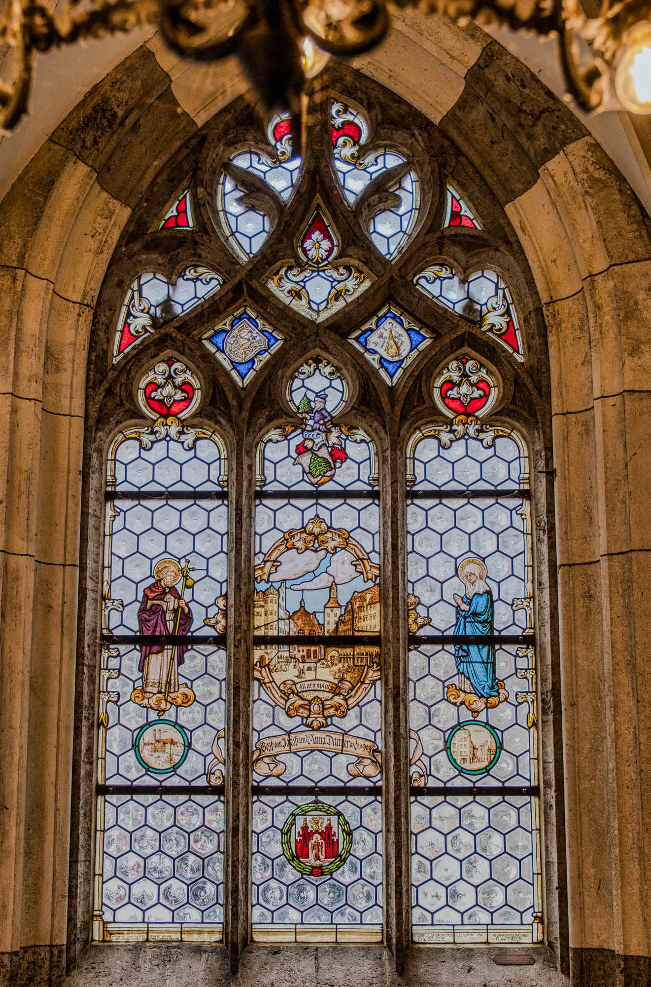 Stained glass in Neues Rathaus (new town hall). It depicts a scenes of historic Munich town center, with a male and female figure on each side with halos behind their head.