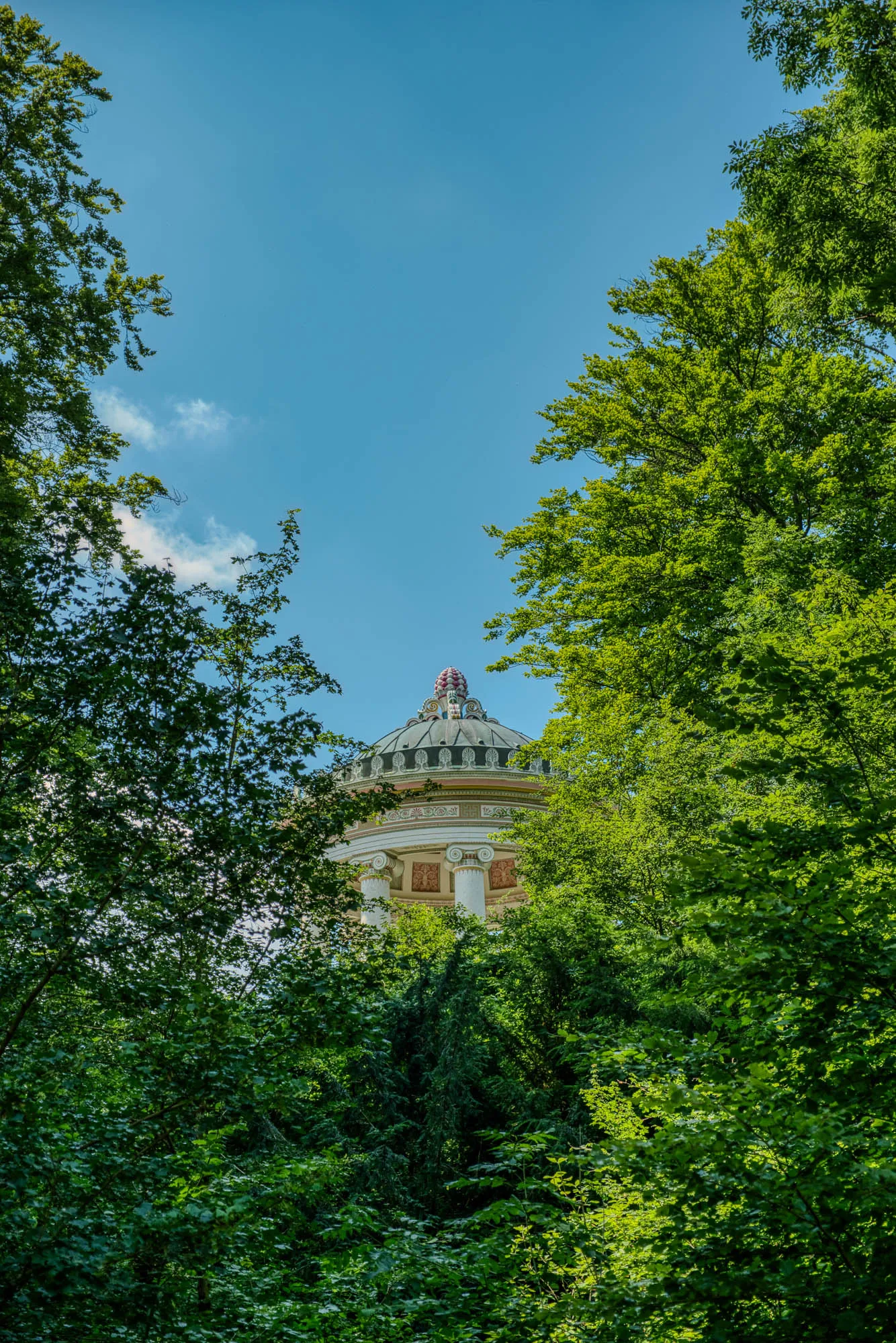 The Monopteros, a Greek-style temple in the English Garden in Munich, peeking through the trees in the foreground, with a light blue sky in the background