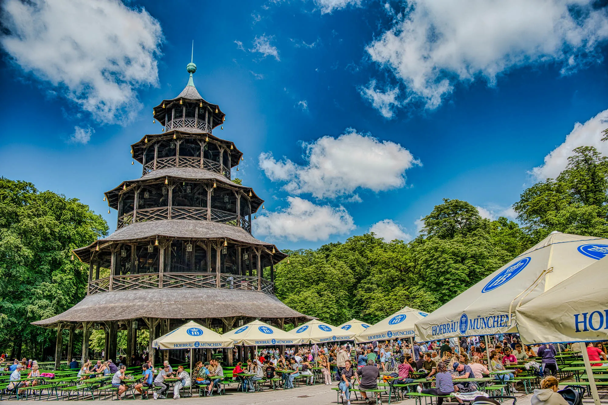 The Chinese Tower, a building resembling a pagoda, and surrounding beer garden in the English Garden in Munich. The beer garden is full of people, there are green trees in the background, and the blue sky has scattered clouds.