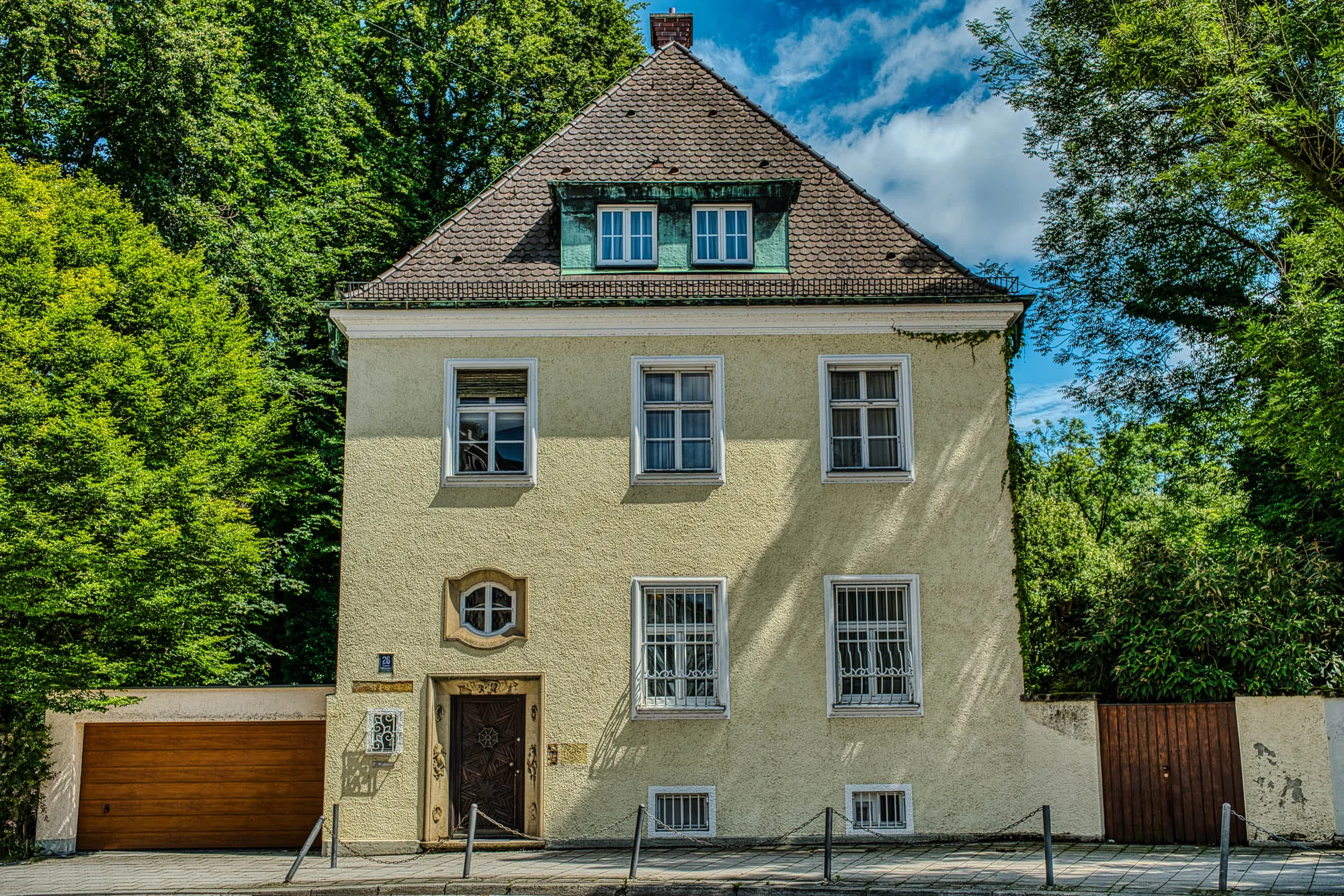 An almost square light yellow house, face on, with a triangular roof and chimney  face-on in fron of a forrest of trees.