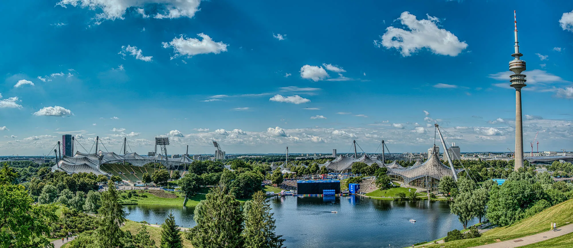 Panorama of Olympic Park in Munich. Olympic tower, hall, and stadium are visible, from right to left. The river is in the foreground, and the fan viewing area for Euro 2024 filled with patrons is in the center.