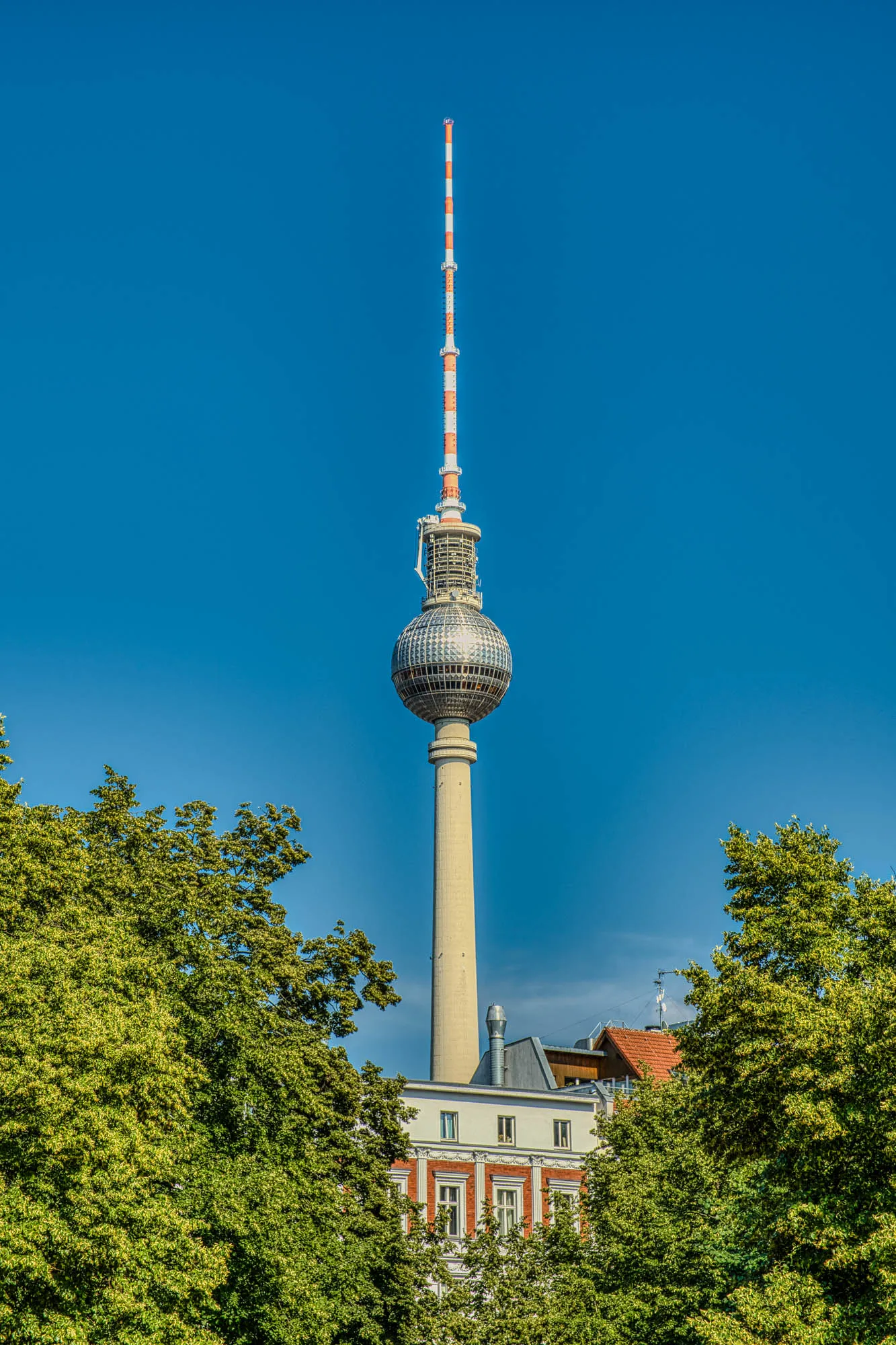 Fernsehturm Berlin appearing over a building in the foreground with green treetops in the foreground. Taken at golden hour with a clear blue sky.