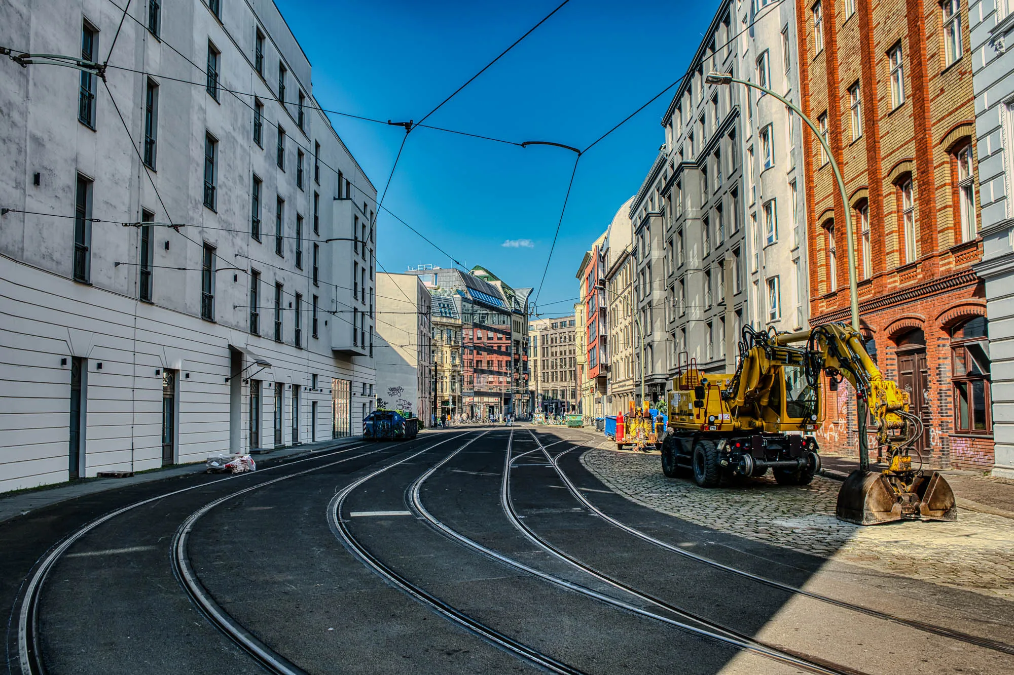 Empty street with cable car tracks in the road and power lines running betwee the buildings. a A digger is parked on the right. At the end of the street a newer all-glass building reflects the older buildings across the street.